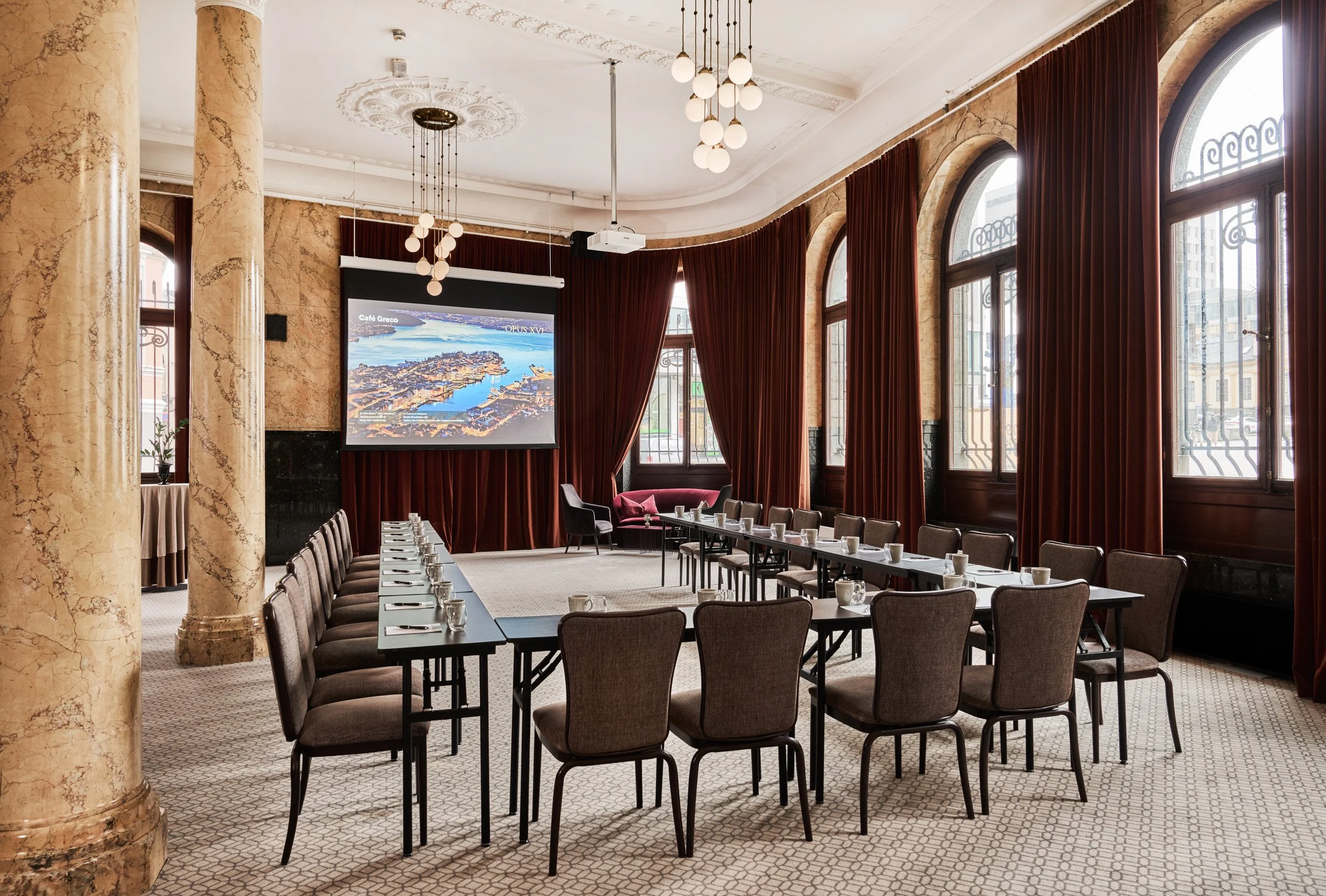 Conference room with U-shaped arrangement of tables and chairs, a large screen, tall arched windows with red curtains, and decorative columns.