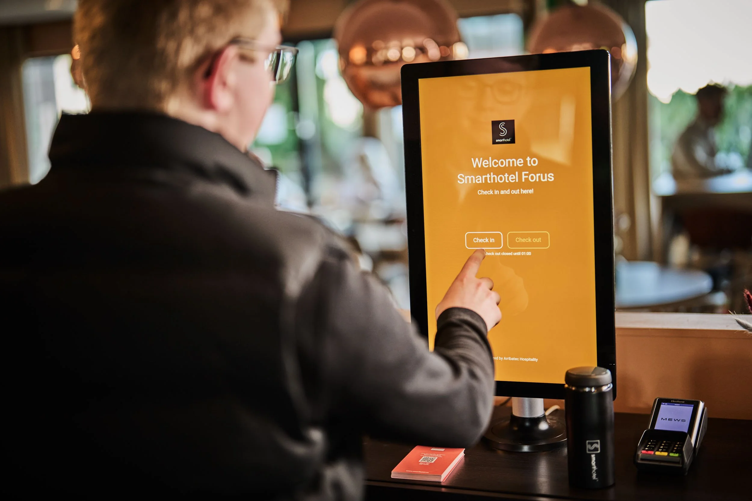 Person in a leather jacket using a touchscreen check-in kiosk at a hotel, with a small black thermos and card on the table.