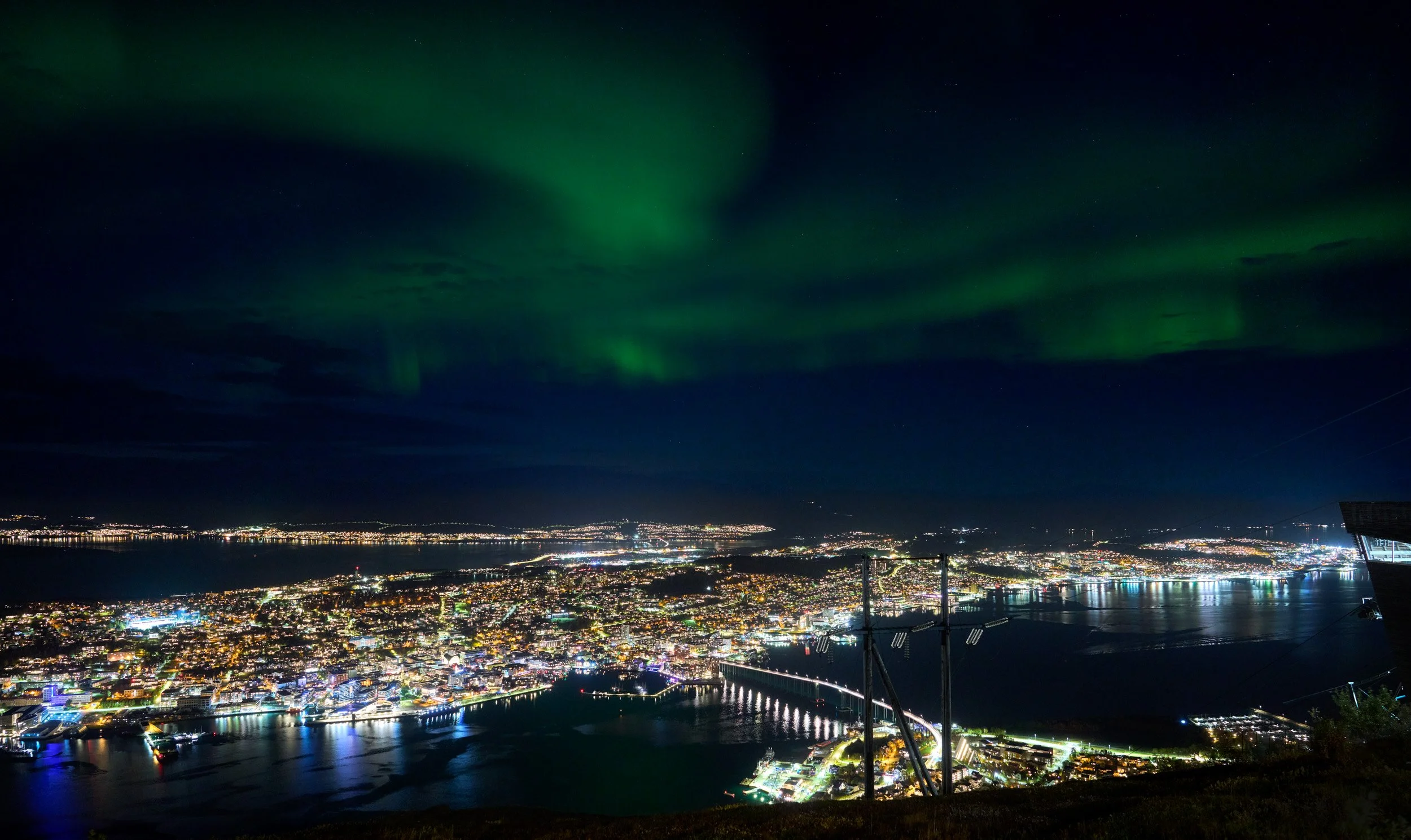 Nighttime cityscape with illuminated buildings and streets, a water body reflecting city lights, and green aurora borealis in the sky.