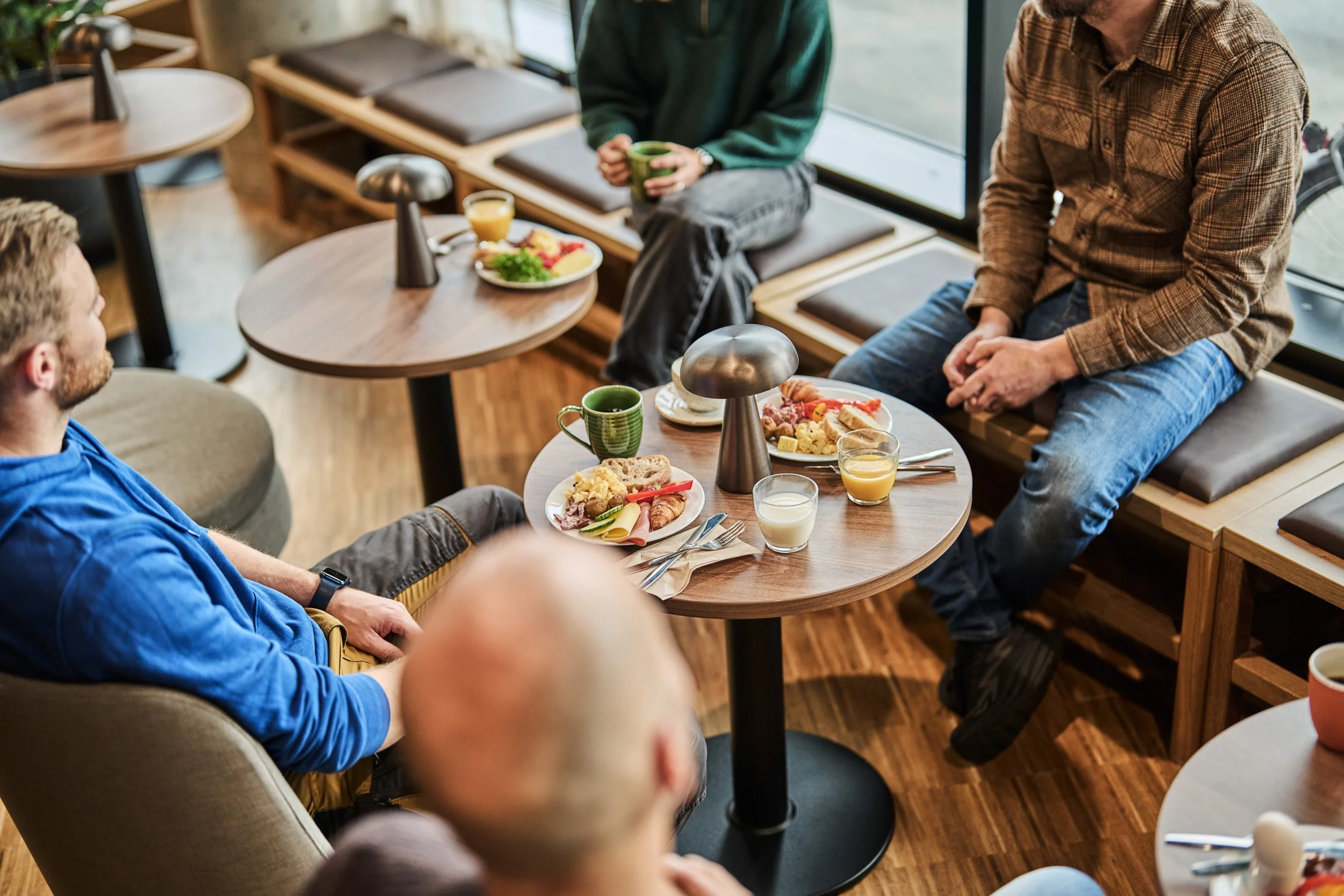 Group of people having breakfast in a cozy cafe with wooden floors and tables, enjoying coffee, juice, and plates of food.