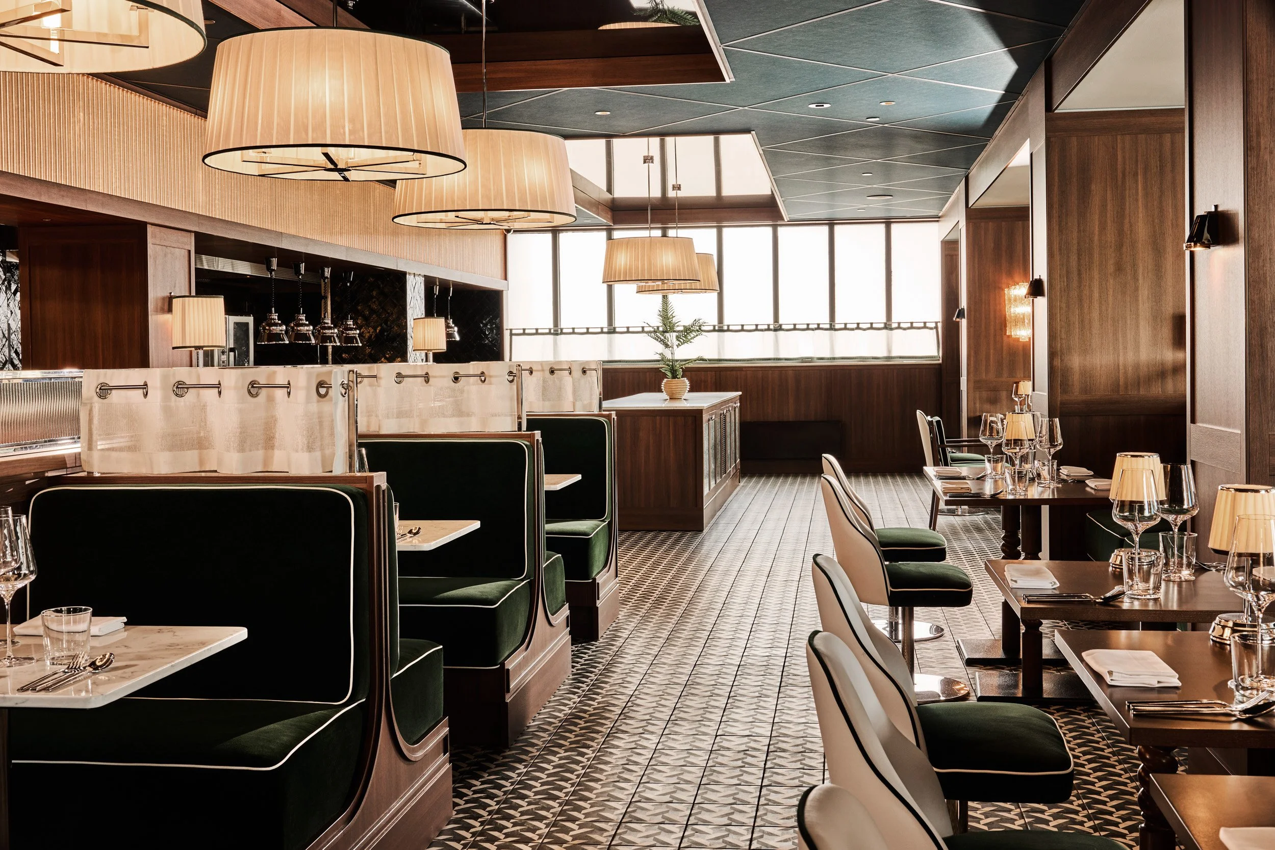 Interior of a restaurant dining area with dark wood paneling, green and white upholstered chairs, arranged for service, with table settings. Large windows with natural light, pendant lights hanging from the ceiling, and a patterned floor.