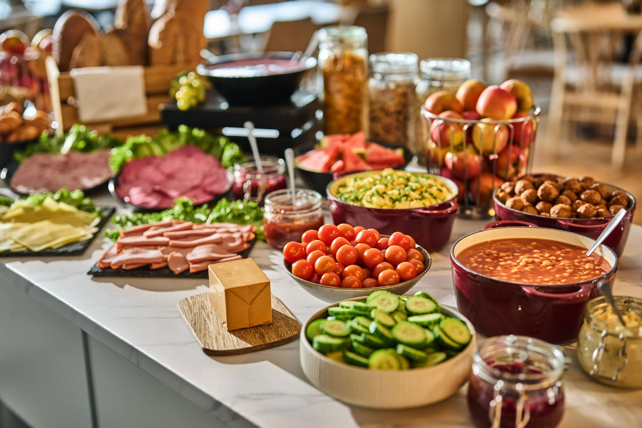 A breakfast or brunch spread on a table with various fresh fruits, vegetables, and breakfast foods including cherry tomatoes, cucumbers, watermelon, apples, grapes, scrambled eggs, bacon, ham, pancakes, nuts, beans, and jams.