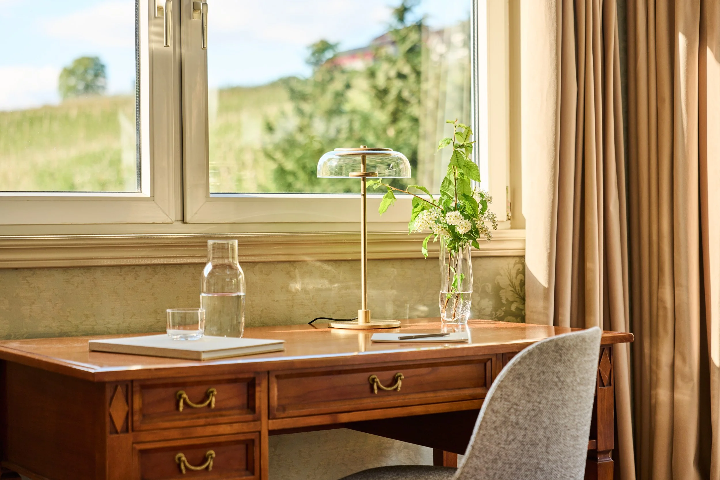 A wooden desk with a glass pitcher of water, a glass, a book, a tall modern table lamp, and a vase with green leaves and white flowers, situated in front of a window with beige curtains, outside greenery visible.