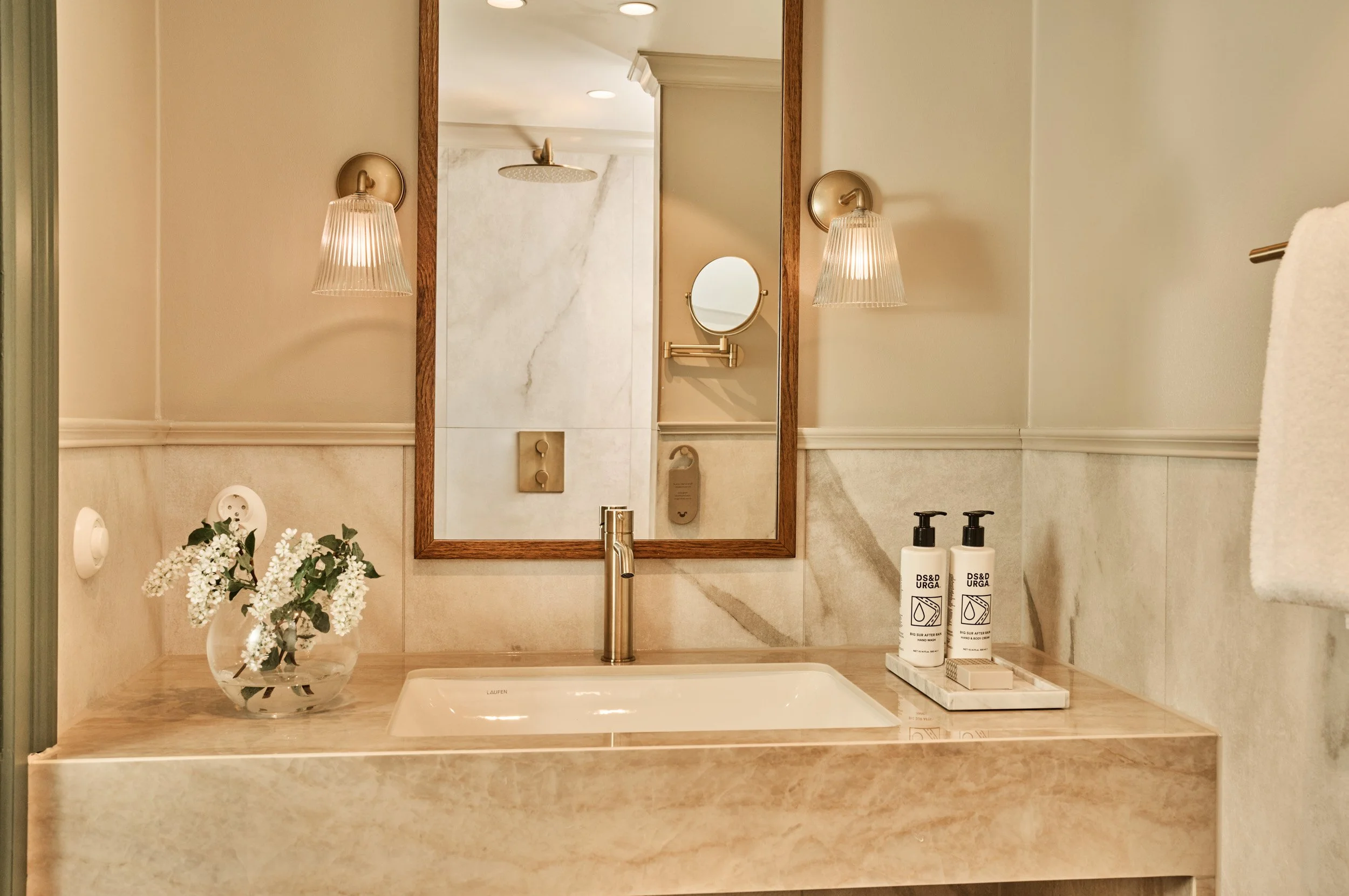 A bathroom at Hotel Ullensvang with a marble countertop, a rectangular sink, a gold faucet, a clear glass vase with white flowers, two soap or lotion bottles, a mirror, wall-mounted lights, and a glimpse of a shower in the background.