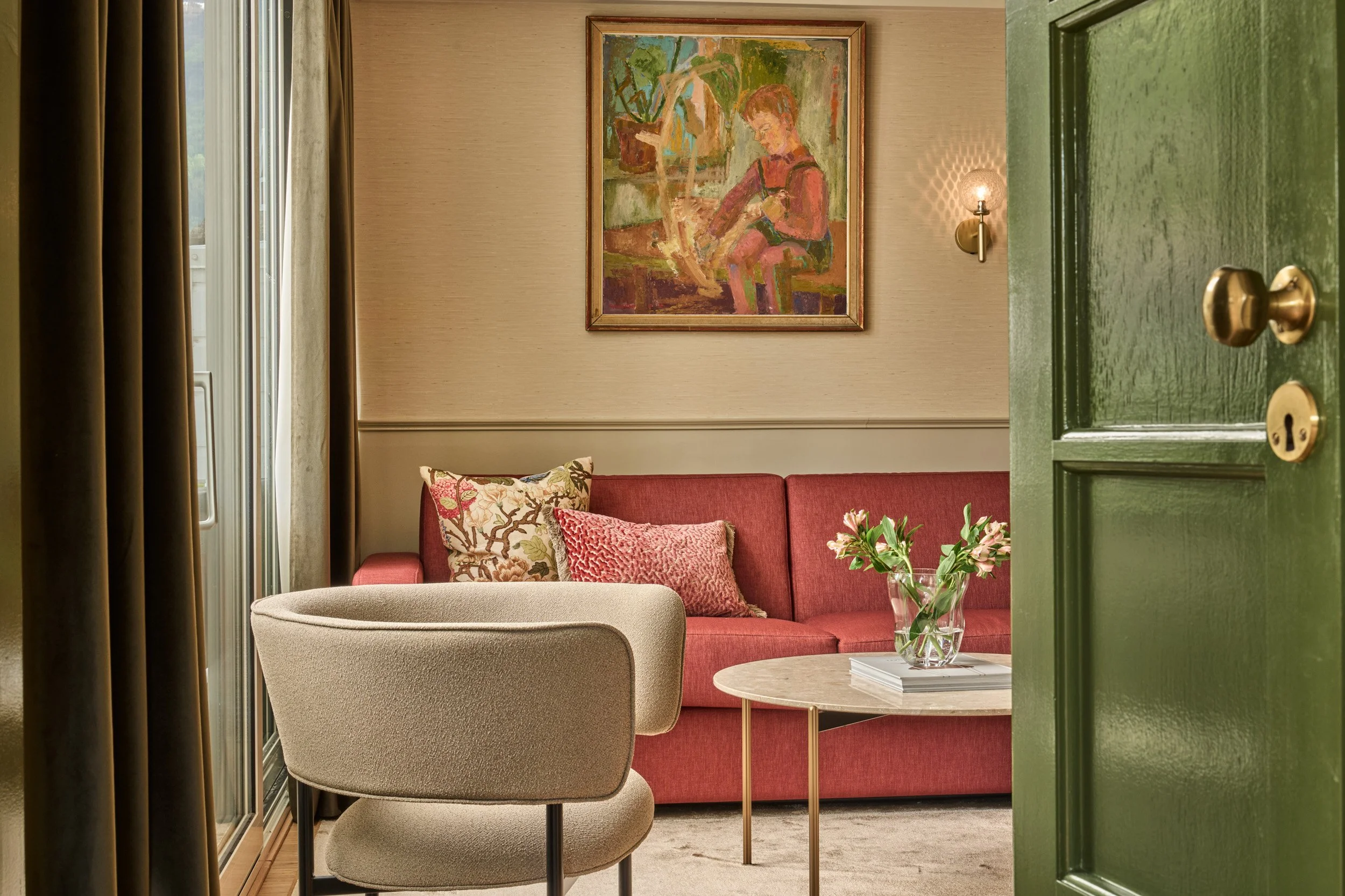 A cozy living room with a red sofa, patterned and textured pillows, a beige armchair, a round marble-topped coffee table with a glass vase of flowers, a painting of a person sitting next to a potted plant hanging on the wall, and a partially open gre