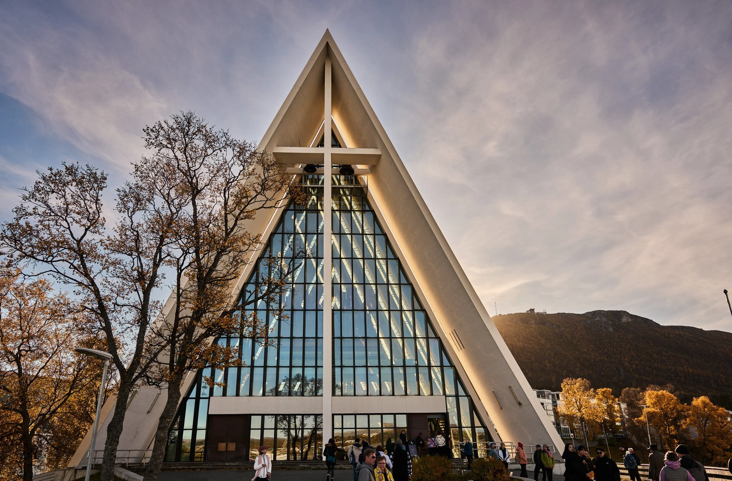 The photo shows an modern A-frame church with a large glass front, surrounded by trees with fall foliage and people gathered outside.