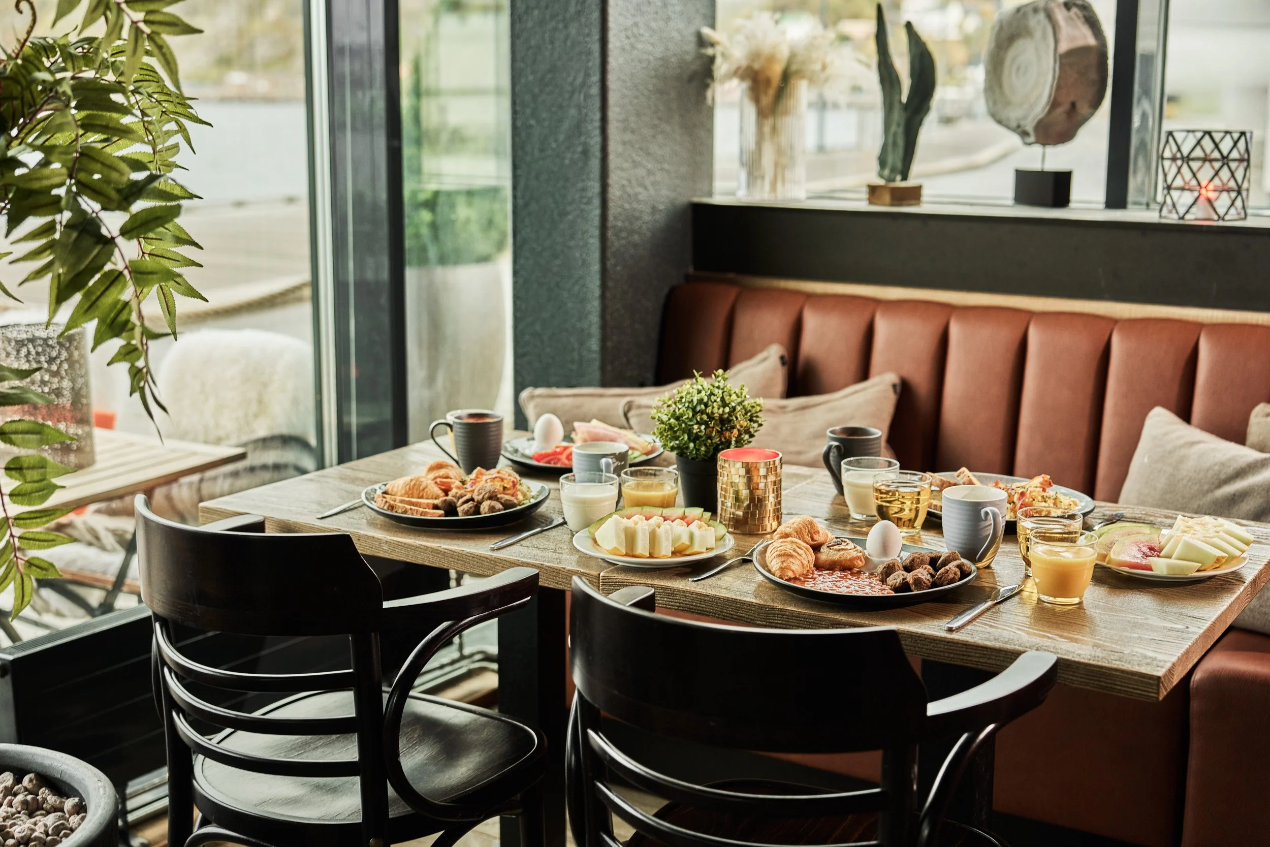 A breakfast table with various foods including pastries, cheese, fruit, eggs, drinks, and condiments in a cozy restaurant or cafe with natural lighting from large windows.