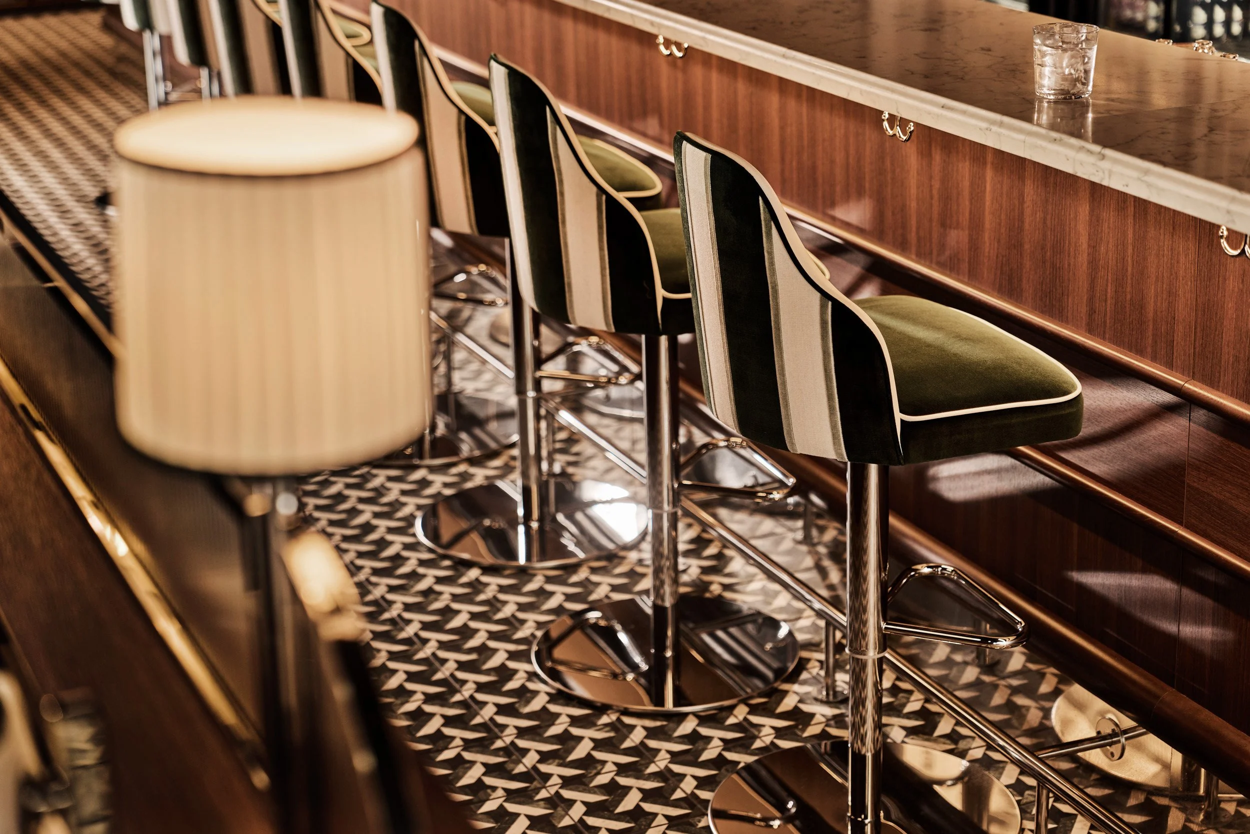 A row of mid-century modern bar stools with green and black striped upholstery, metal bases, and footrests, positioned along a wooden bar counter with a marble top, in a vintage-style bar or lounge with patterned carpet flooring.