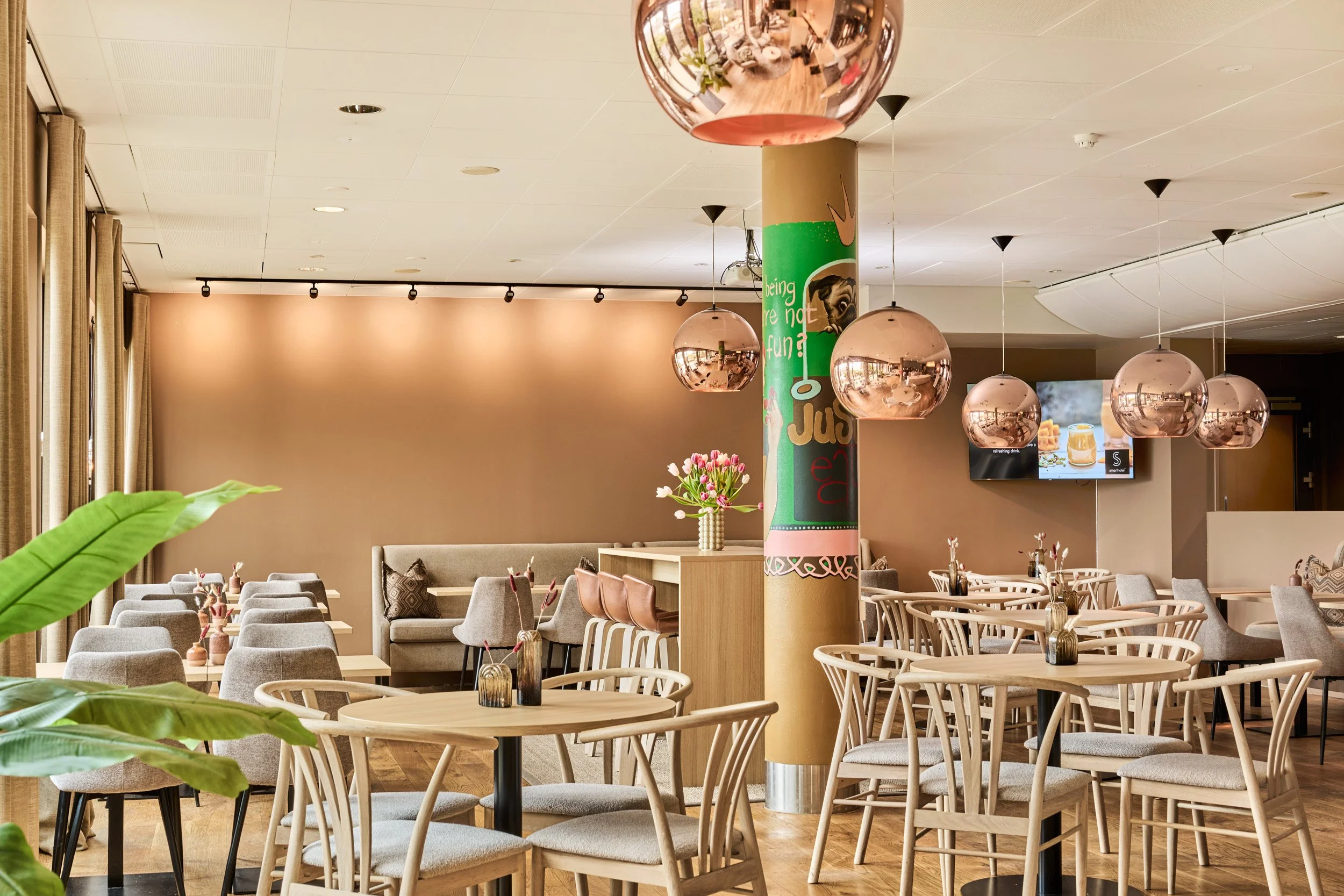 Empty modern restaurant with round and rectangular tables, gray and wooden chairs, pink pendant lights, a floral arrangement, large window with curtains, and a TV screen showing a drink