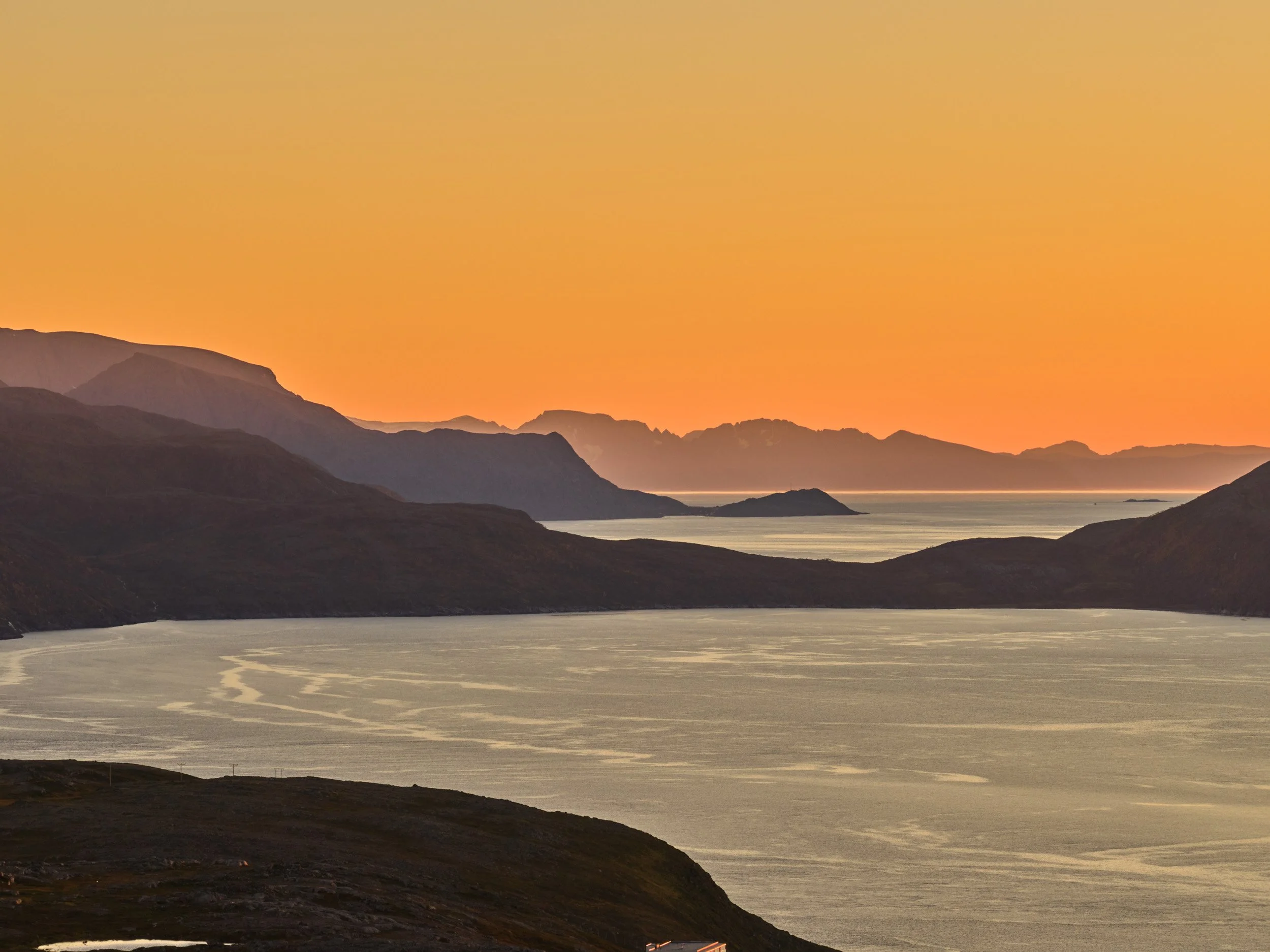 Sunset over a mountainous landscape with a body of water in the foreground and distant islands.