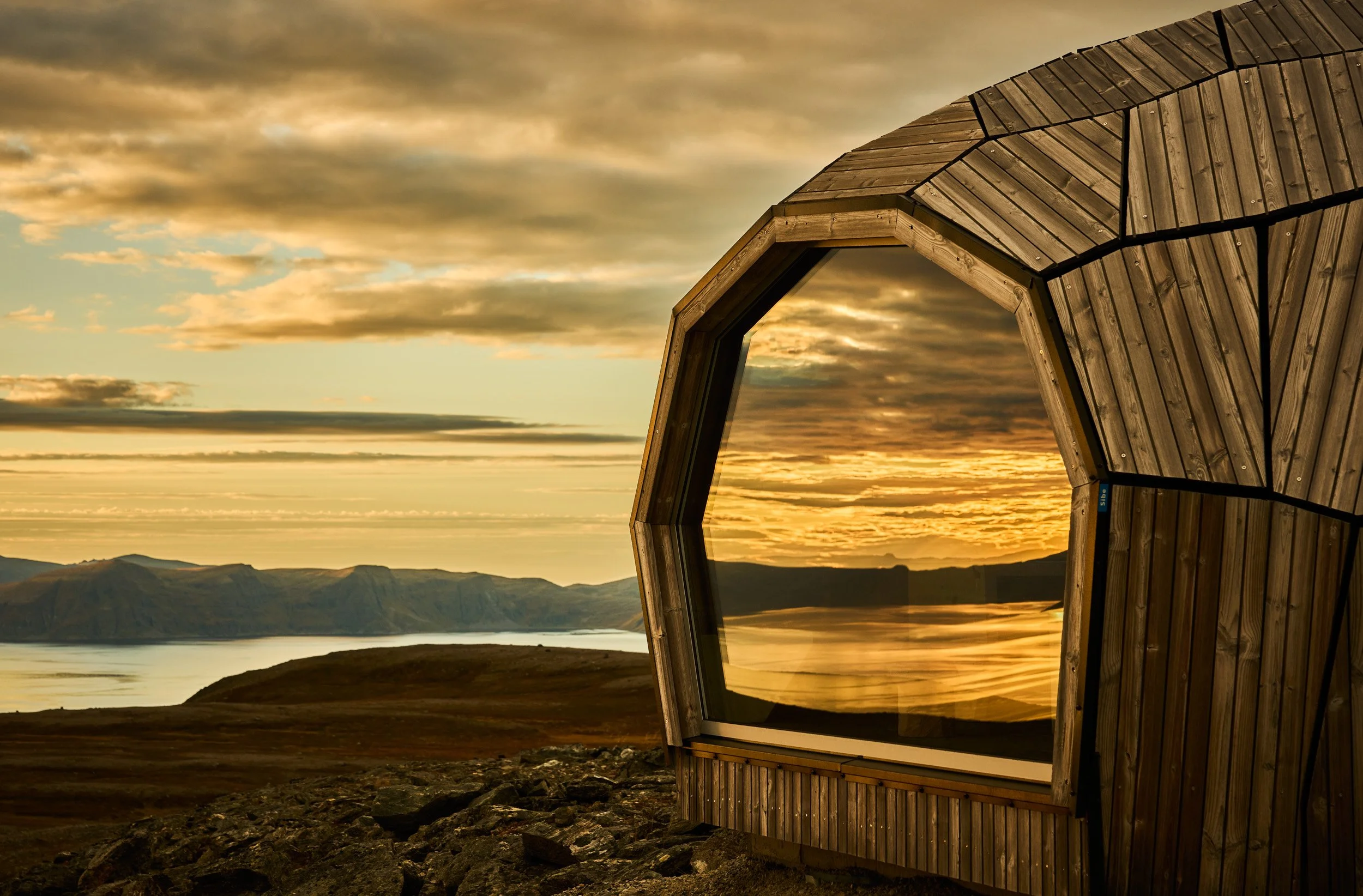 A wooden structure with an irregular shape and large window, positioned outdoors on a rocky terrain at sunset or sunrise, overlooking rolling hills, a body of water, and mountains in the distance.