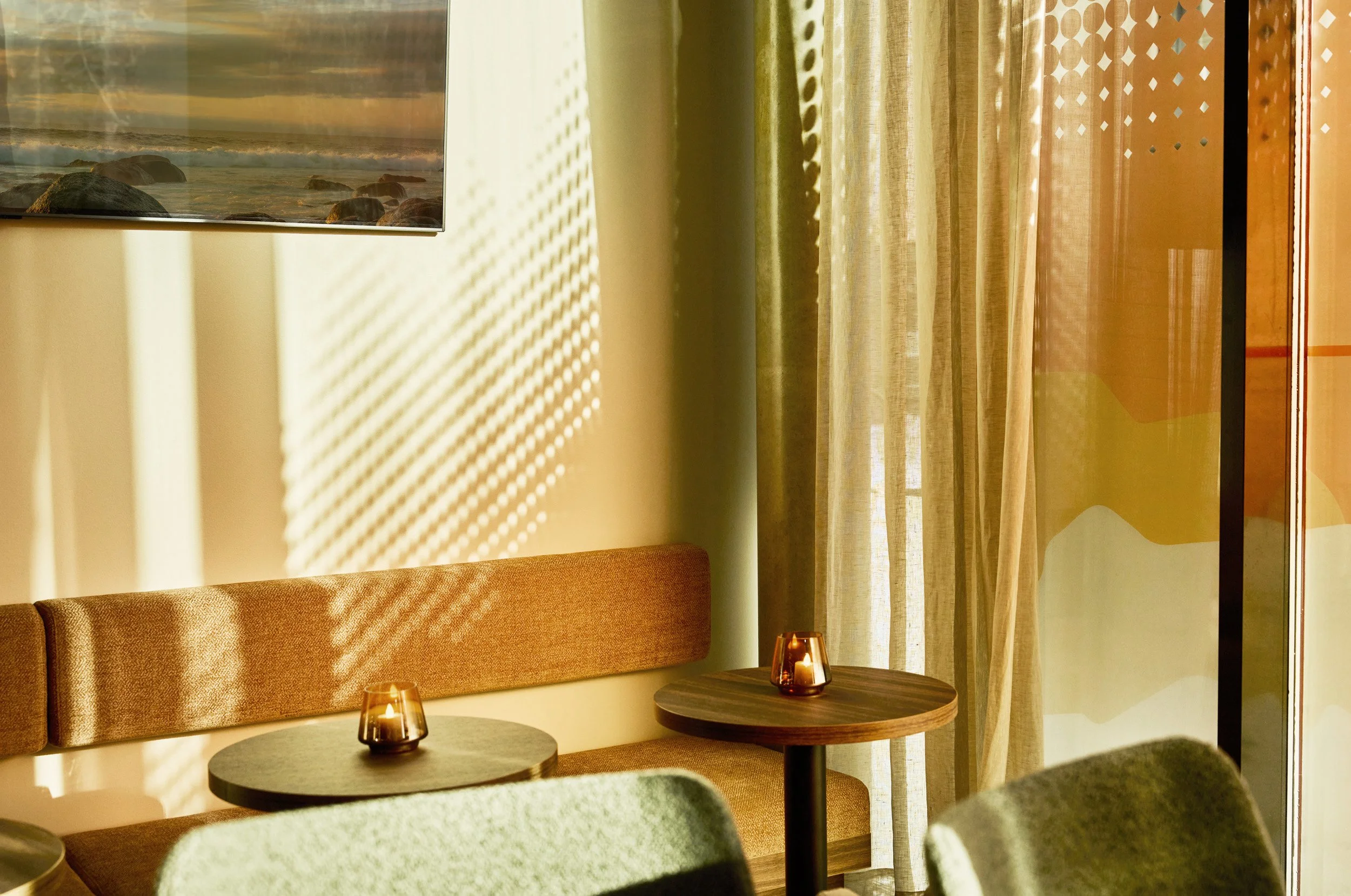 A cozy corner in a room with a beige cushioned bench, two small round wooden tables, and candles inside glass holders on each table. Light filters through curtains, casting shadows on the wall, with a portion of a painting visible in the top left cor