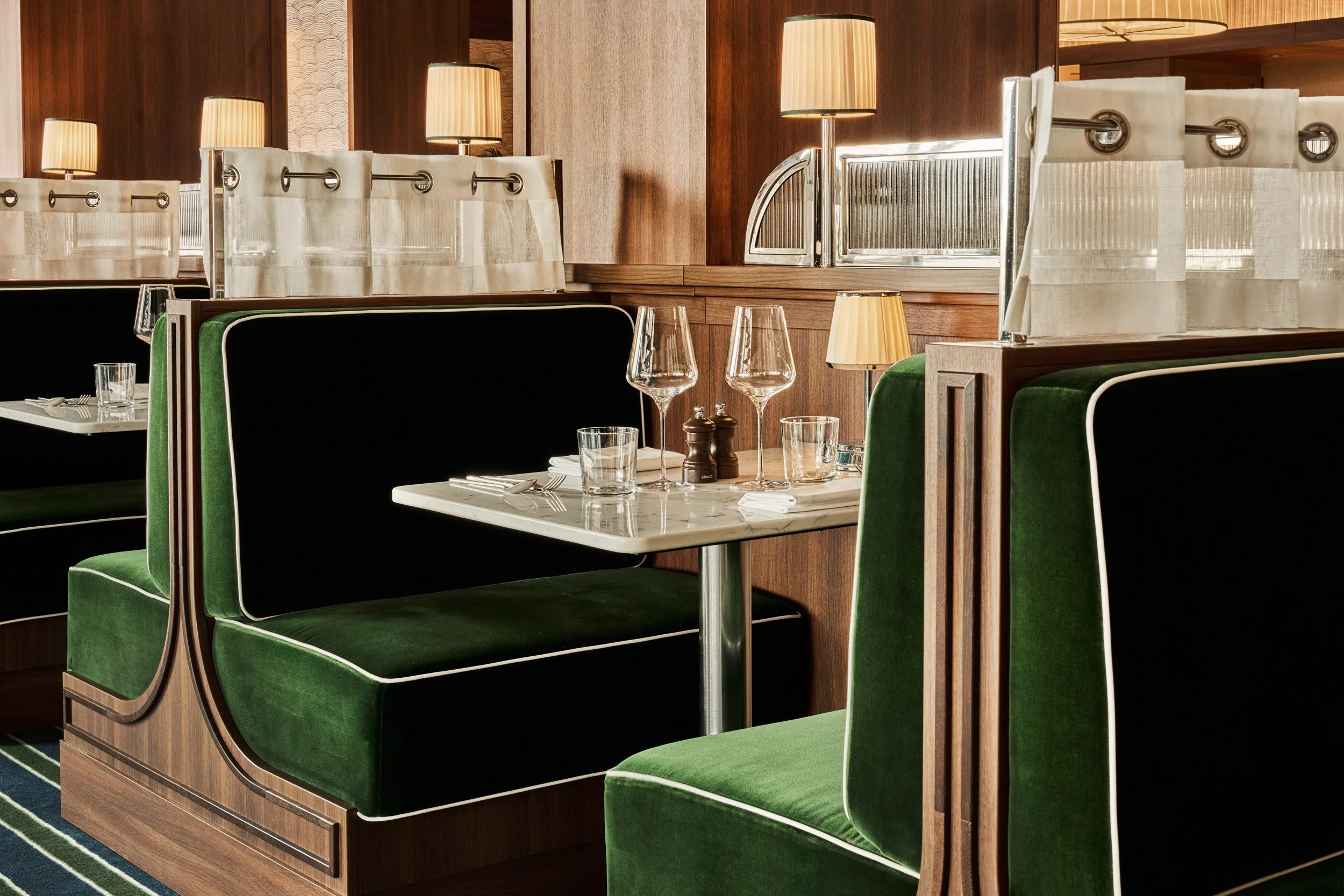 A cozy restaurant booth with green upholstered seats, a marble table set with glasses, salt and pepper shakers, and white napkins, illuminated by warm lighting and wooden paneling.