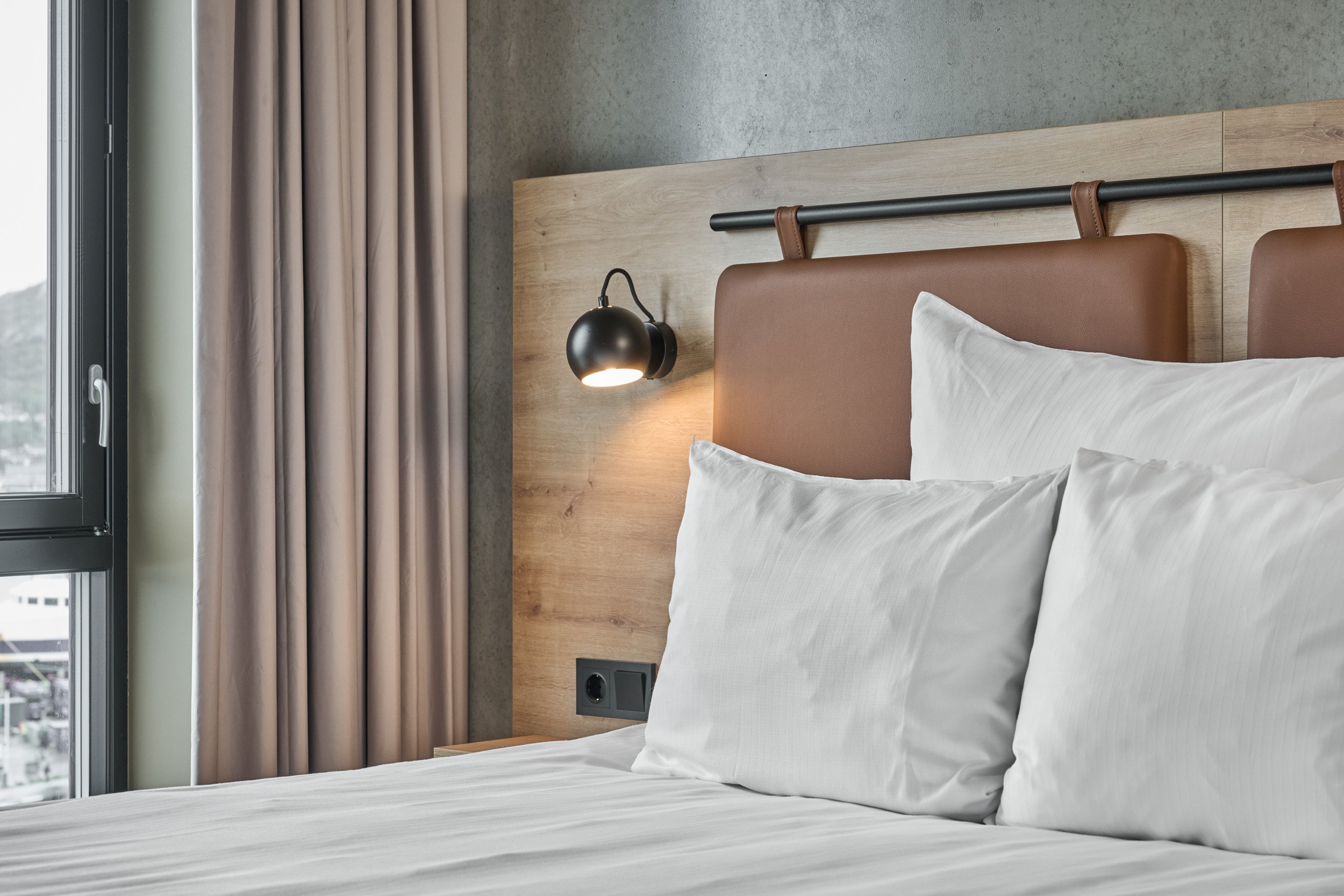A neatly made bed with white pillows and sheets, positioned next to a large window with beige curtains, in a modern hotel room with a wooden headboard and a black wall-mounted reading light.