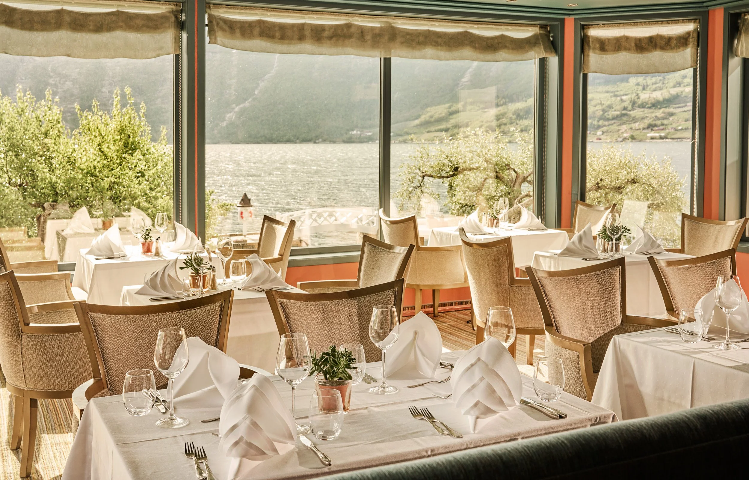 A restaurant with large windows overlooking a lake and green mountains, set with white tablecloths, folded napkins, wine glasses, and small potted plants.