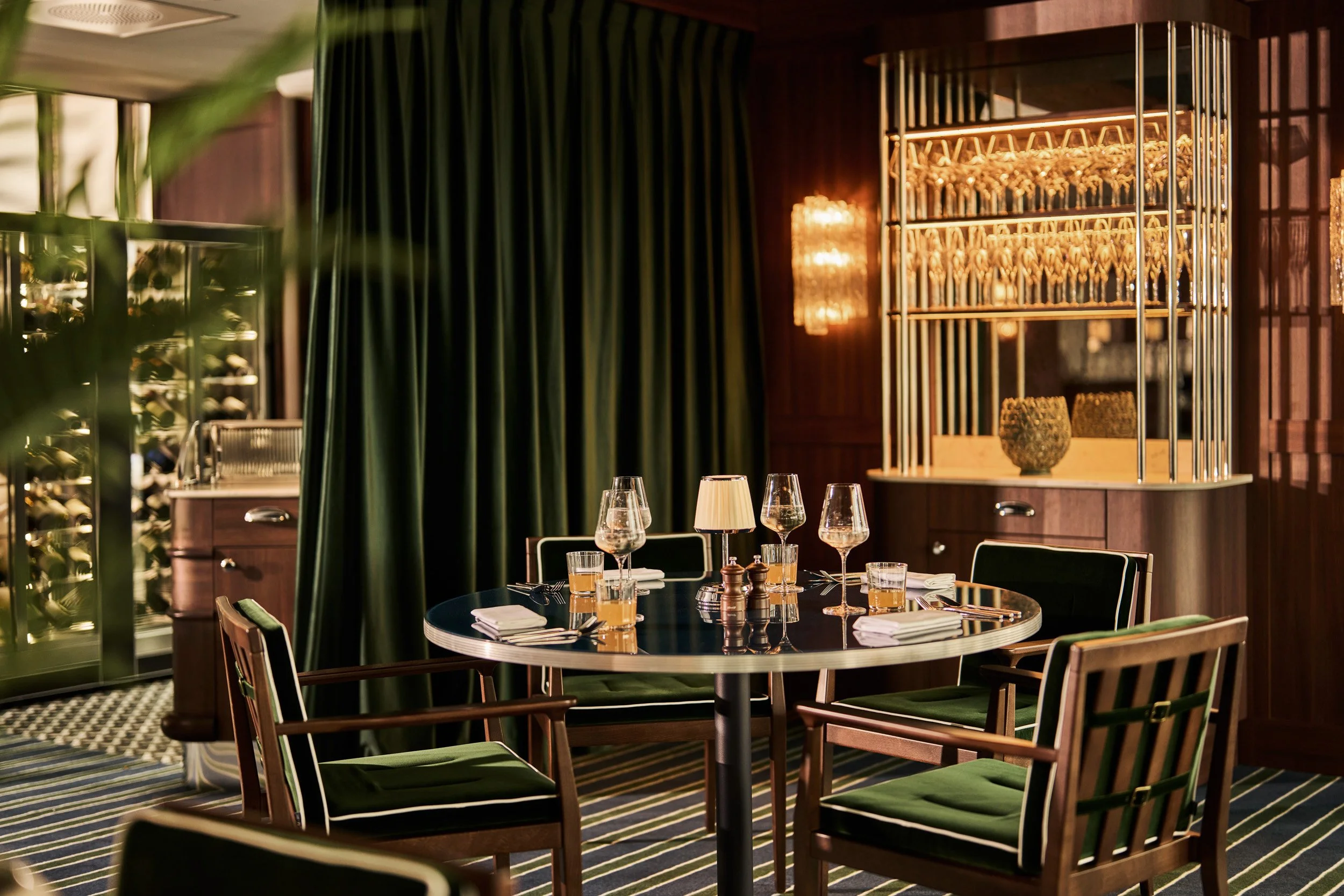Elegant restaurant dining area with a polished round table set with wine glasses, water glasses, napkins, and a small lamp. Surrounding the table are wooden chairs with green cushions. The background features dark green curtains, a wooden cabinet wit