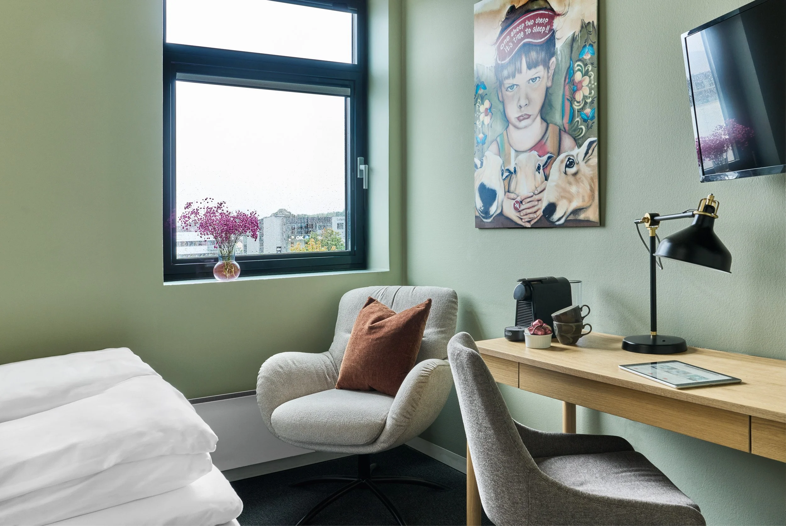 A cozy hotel room with a bed, a fluffy armchair with a brown pillow, a wooden desk with a table lamp, a coffee machine, and cups. A window with a vase of pink flowers on the sill shows a cityscape outside. A colorful portrait of a woman with dogs han