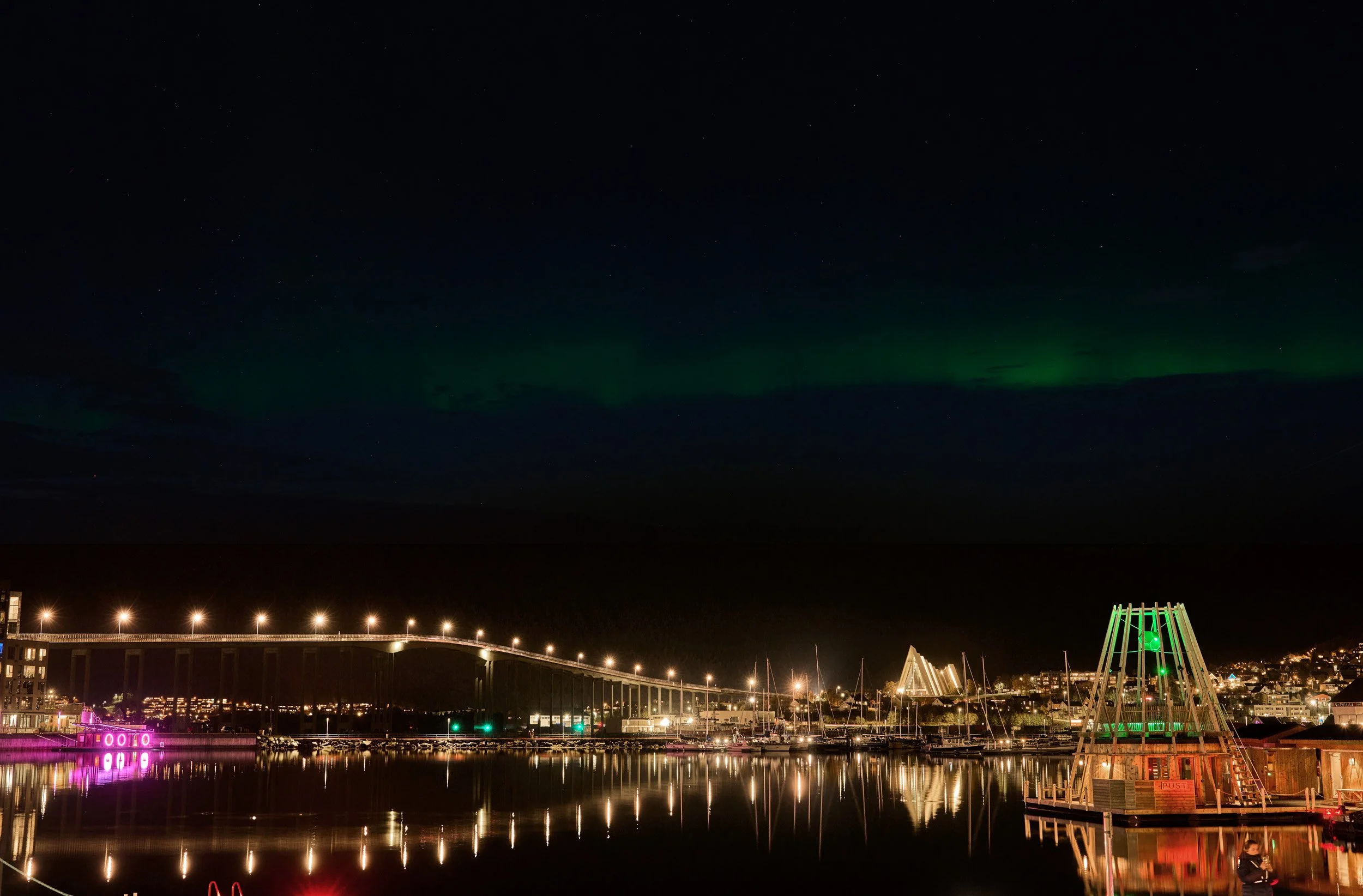 Night scene of a harbor with a bridge and boats, city lights, and an aurora borealis in the sky.