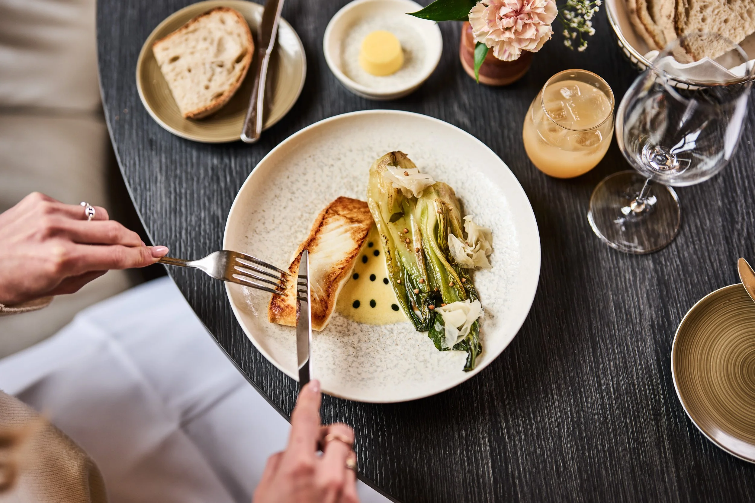 Person using a fork and knife to cut into a piece of fish on a white plate, with grilled greens and a sauce, on a dark dining table. Surrounding table includes bread, butter, pink flowers, and drinks including a glass of white wine and a pink cocktai