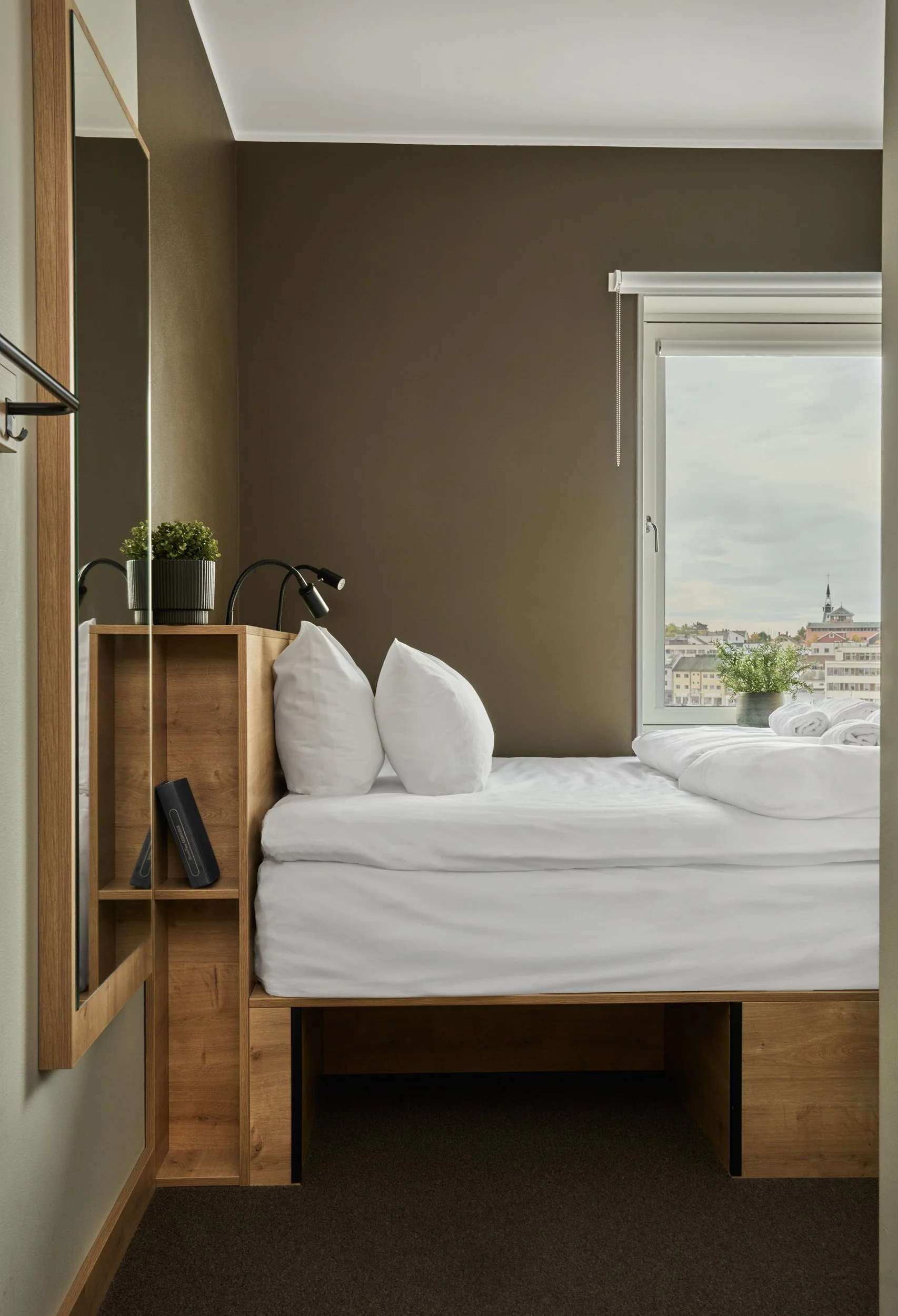 A neatly made bed with white linens and pillows in front of a window showing a cityscape with buildings and a church steeple, with a potted plant on the windowsill and a brown wall.