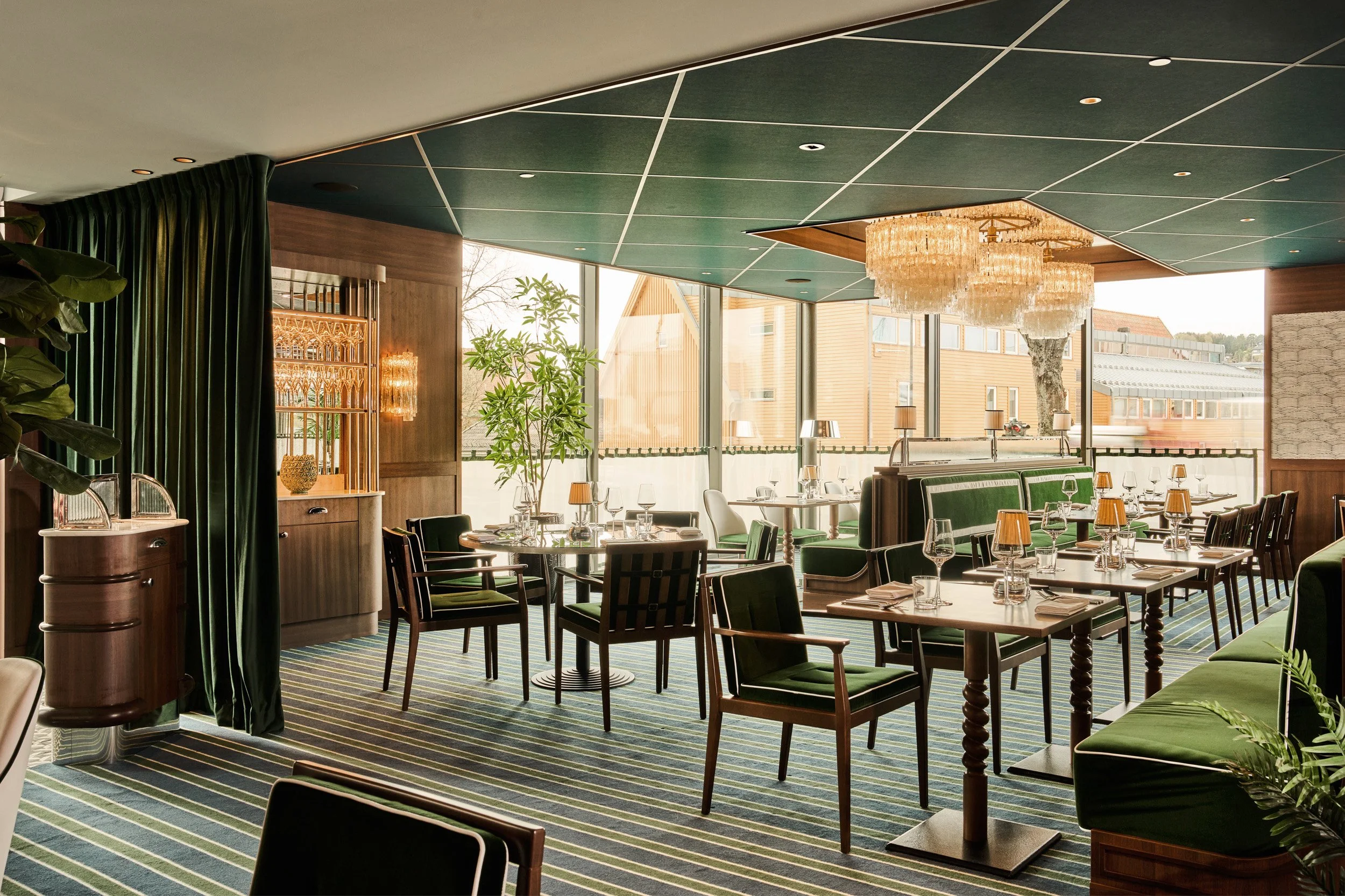 Elegant restaurant interior with wooden and green decor, tables set with glasses, napkins, and small lamps, large windows with natural light, and a chandelier hanging from the ceiling.