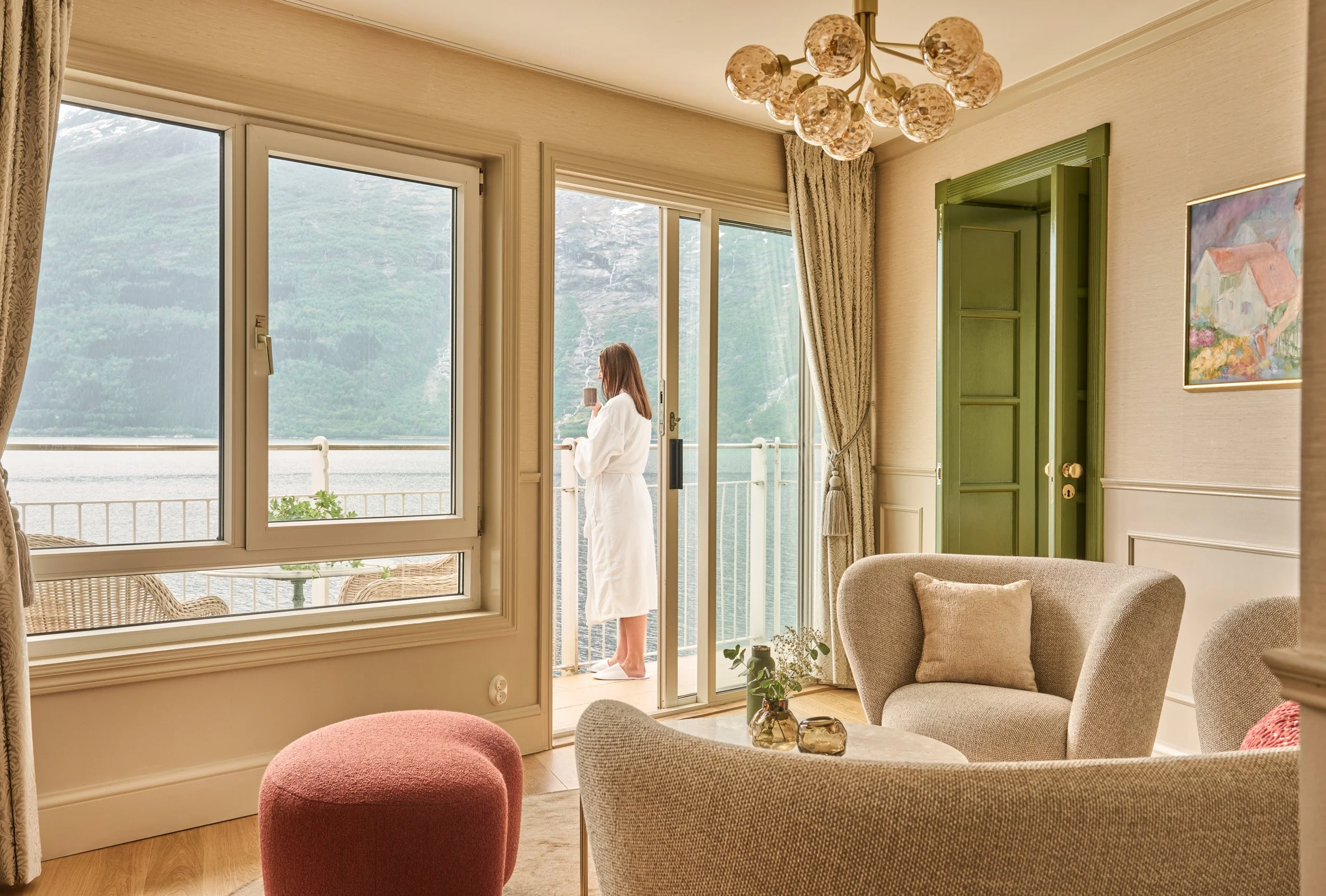 A woman in a white bathrobe standing on a balcony, drinking coffee or tea, with a mountain lake view in the background, inside a cozy living room.