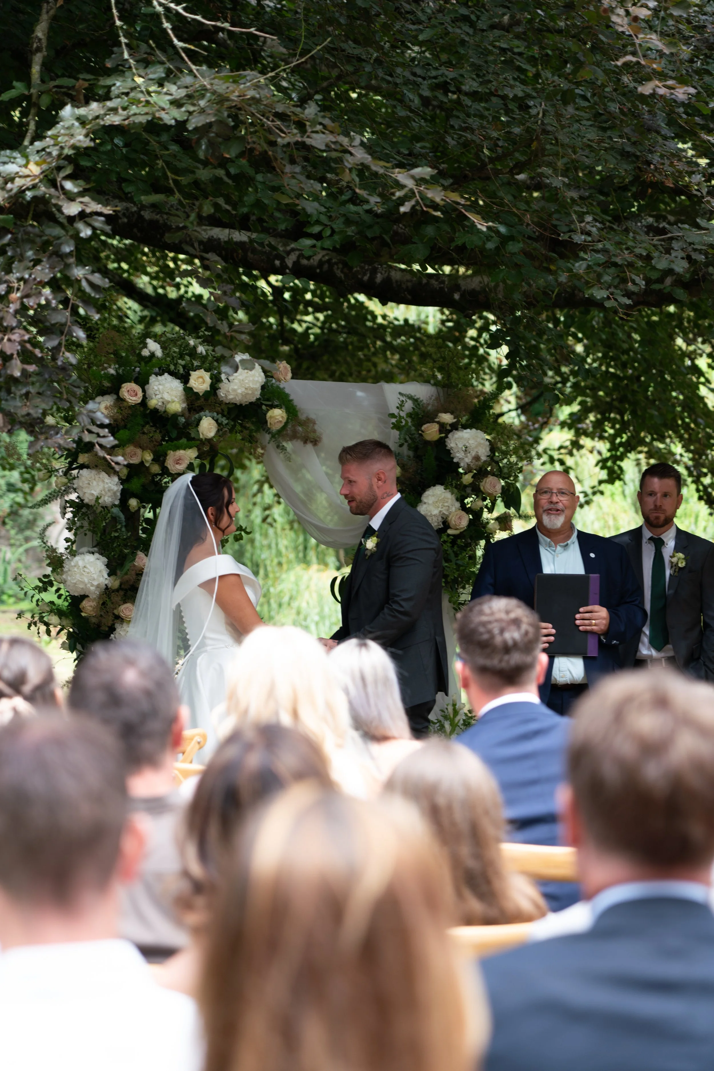 A bride and groom standing face to face at their outdoor wedding ceremony under a canopy of green leaves, exchanging vows, with an officiant holding a book and guests seated in the foreground.