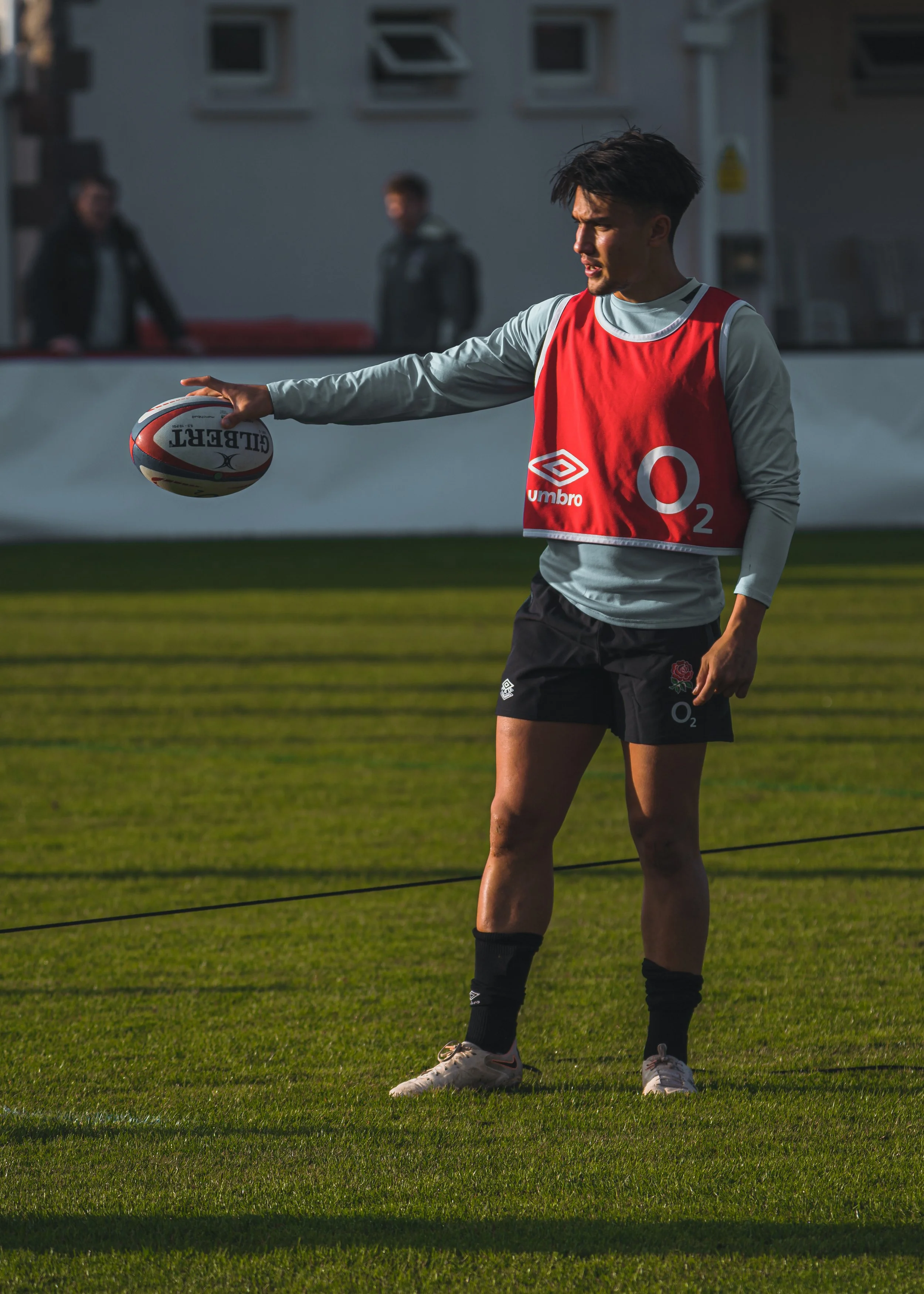 Marcus Smith wearing a red England Rugby training vest holding a rugby ball in a single hand on a grassy field during daytime.
