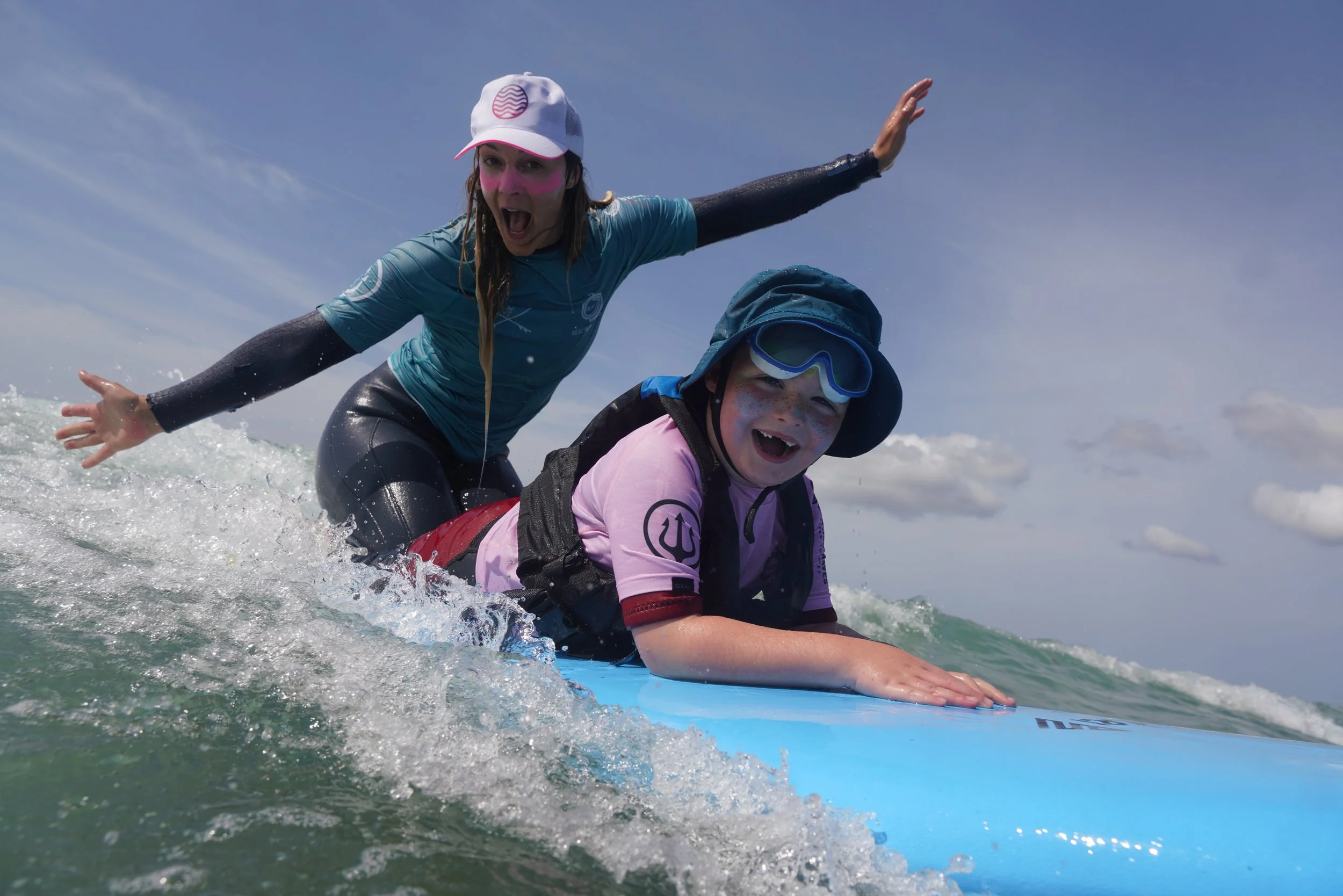 A woman and a young girl are riding a surfboard on the water, with the woman balancing with her arms outstretched, both wearing hats and protective clothing, enjoying a day at the beach with the sky and ocean in the background.