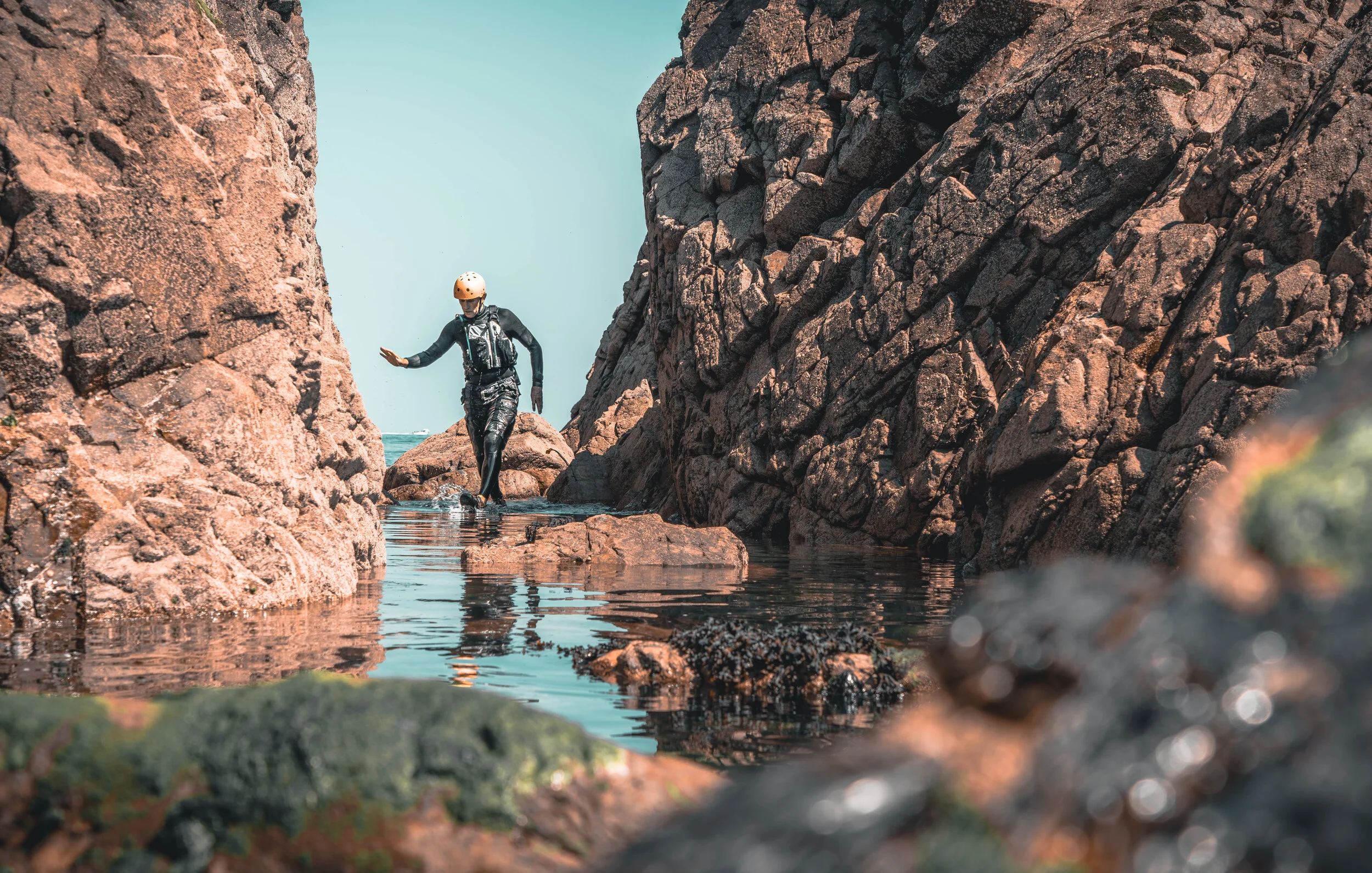 A person in a wetsuit and climbing helmet walking through a narrow rocky canyon with water at their feet.