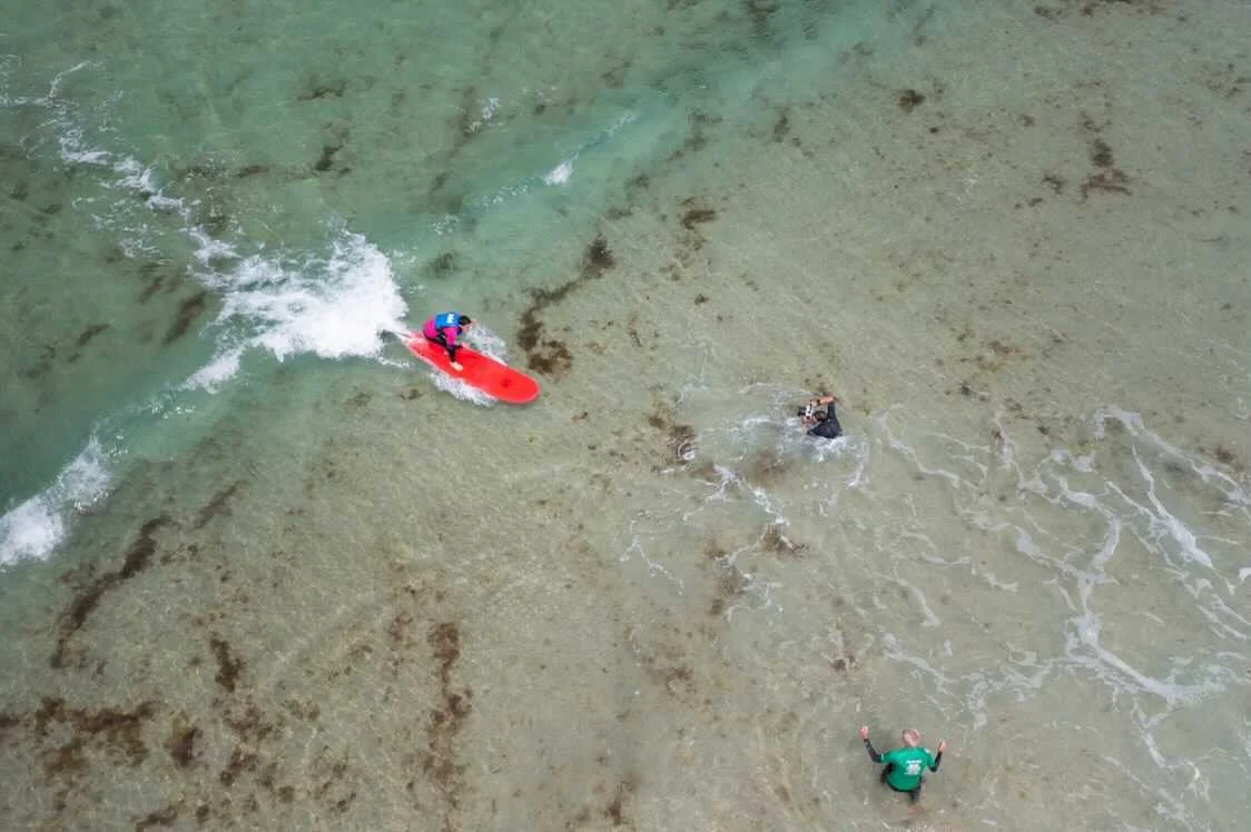 A drone photograph of a person riding a red surfboard, a photographer in dark wetsuit in the water.