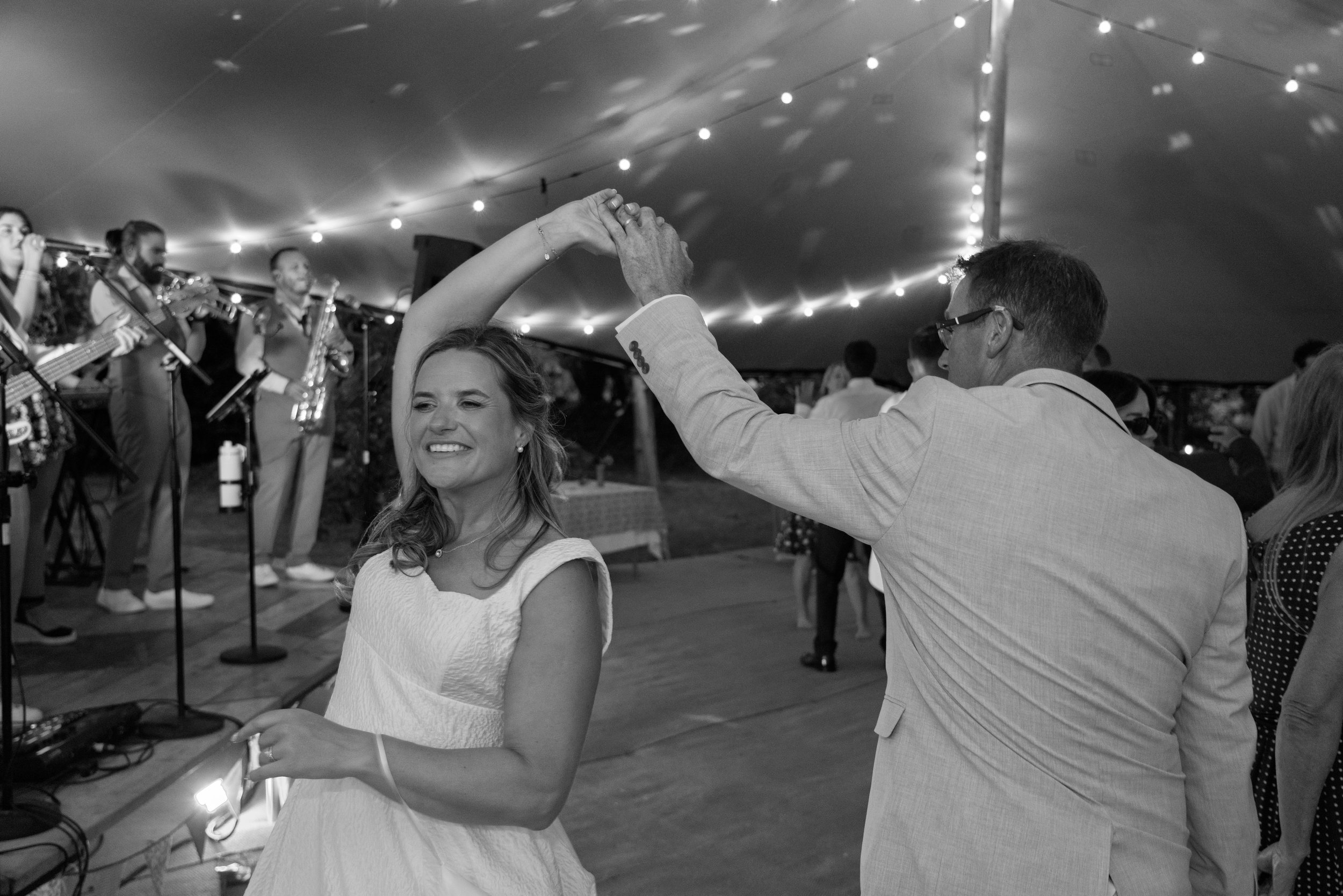 A couple dancing at a celebration, with the background showing a live band playing music under string lights.