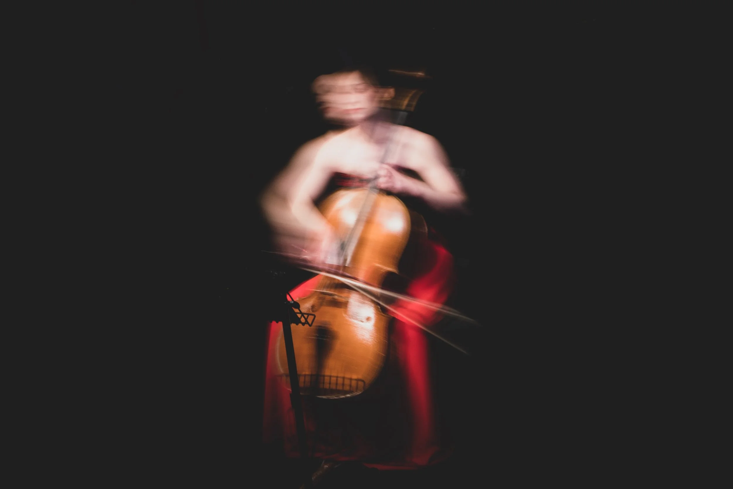 Blurred image of a woman playing cello on a dark stage, wearing a red dress, with a music stand in front of her.