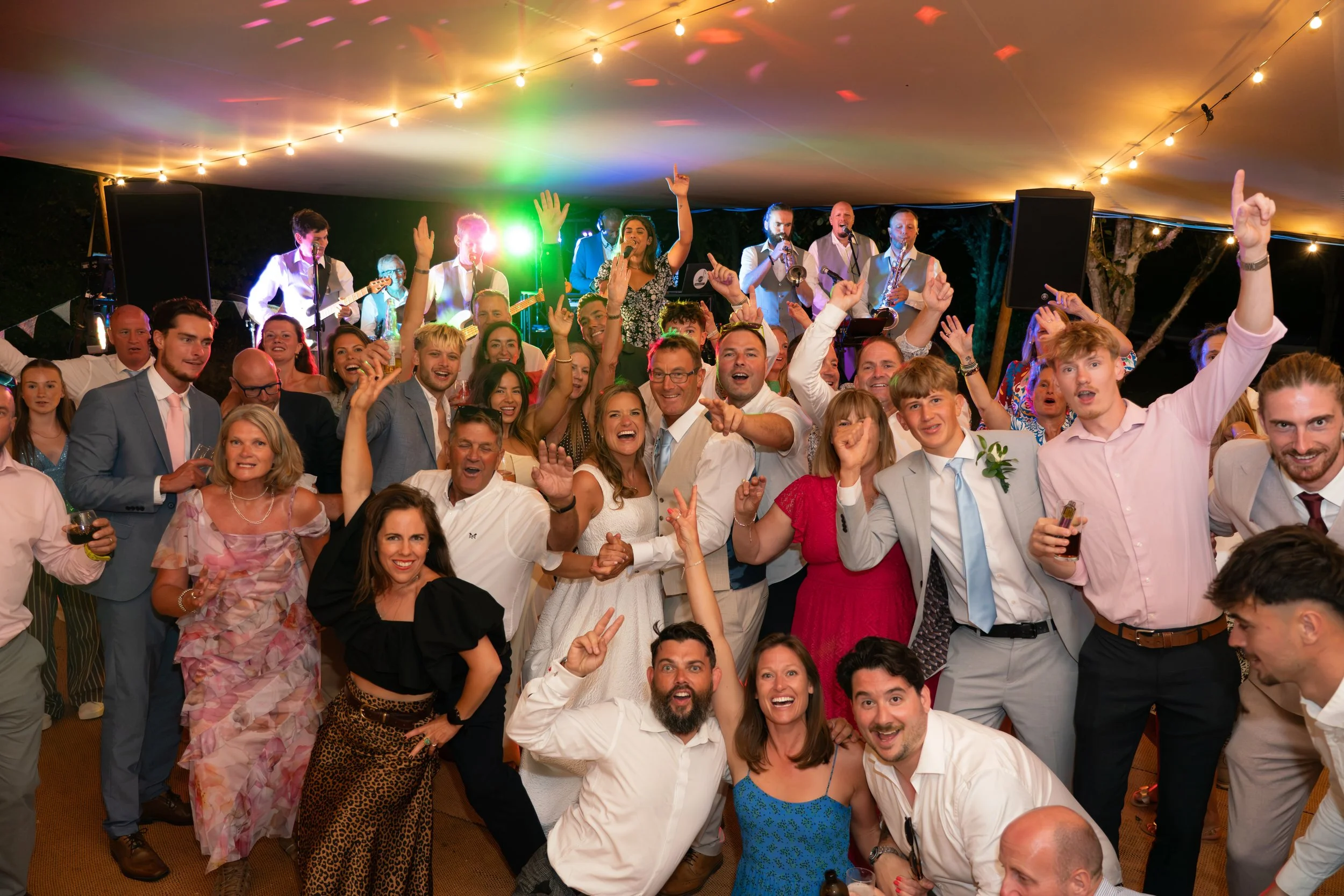 A lively crowd of people at a wedding, dancing and smiling, with a band playing on stage under string lights at night.