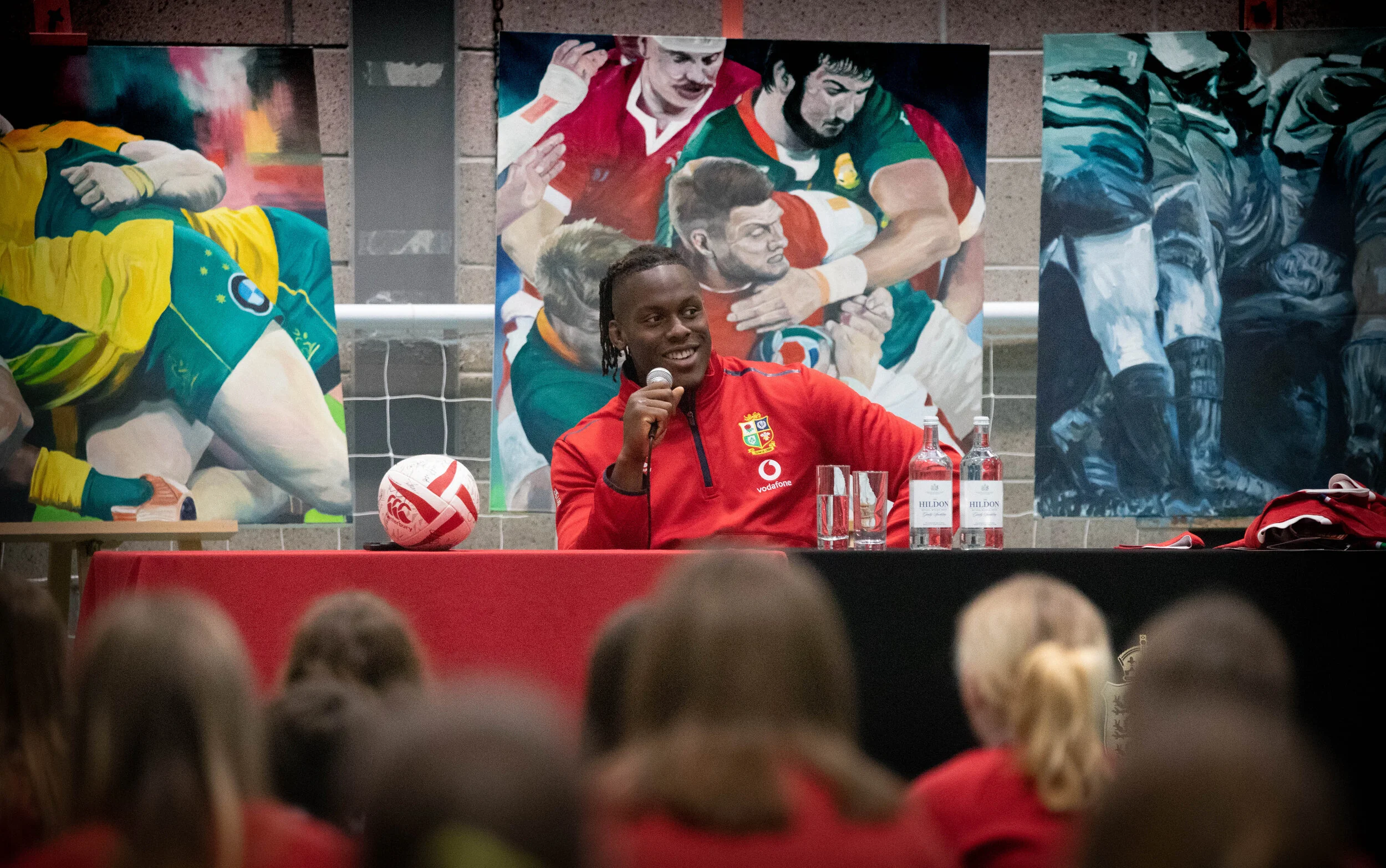 Maro Itoje in a red B&I lions jacket sitting at a table with a microphone, with artworks of rugby players in action hanging on the wall behind him and a group of school children in the foreground.