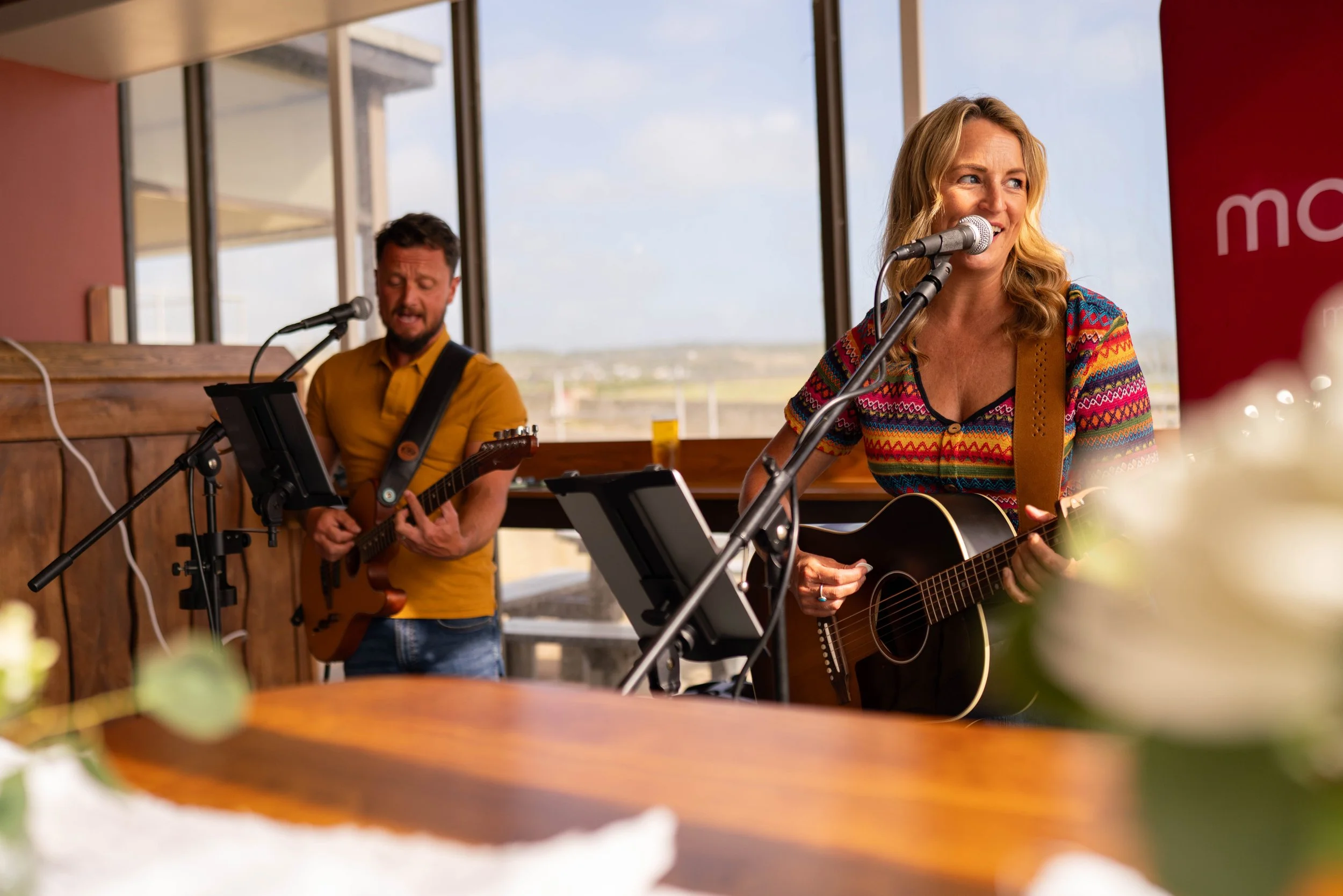 A woman with blonde hair singing and playing an acoustic guitar during a live performance indoors, with a man in the background playing an electric guitar, and a scenic view through large windows.