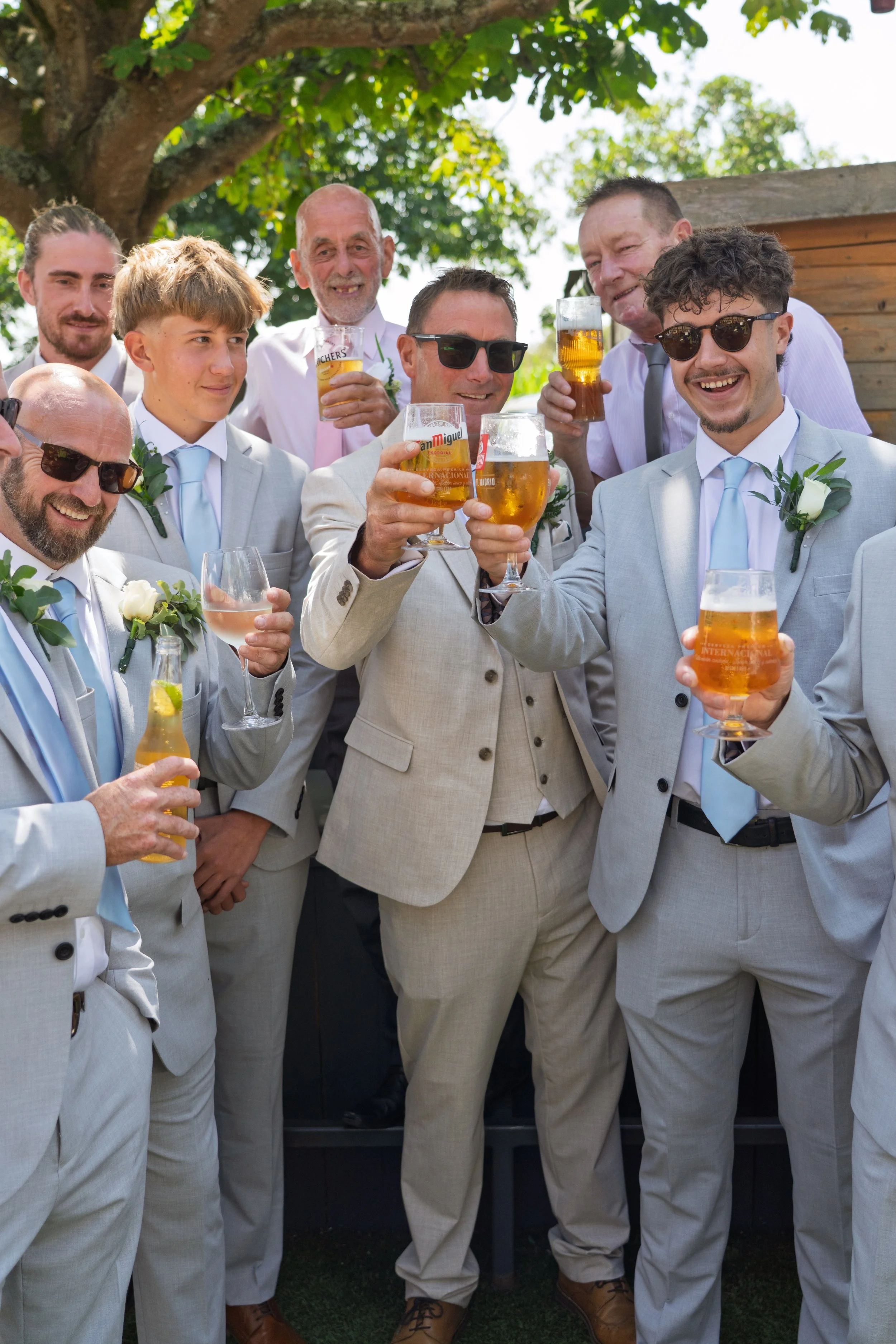 A group of men dressed in light-colored suits celebrating with drinks outdoors under a large tree.