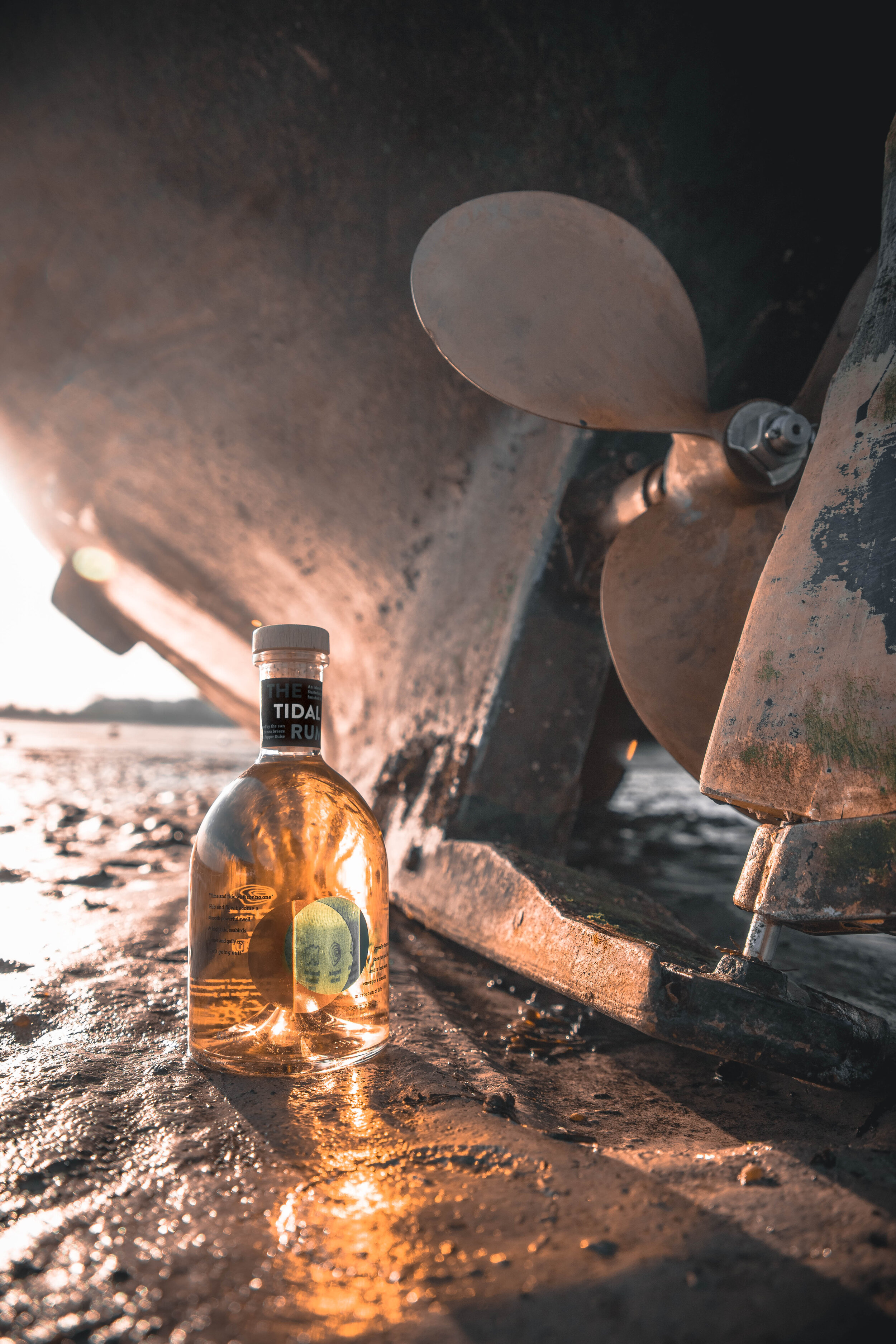 A bottle of alcohol labeled 'The Tidal Run' resting on wet sand near a large, rusted ship's propeller at sunset, with the reflection of the sunset on the bottle and sand.