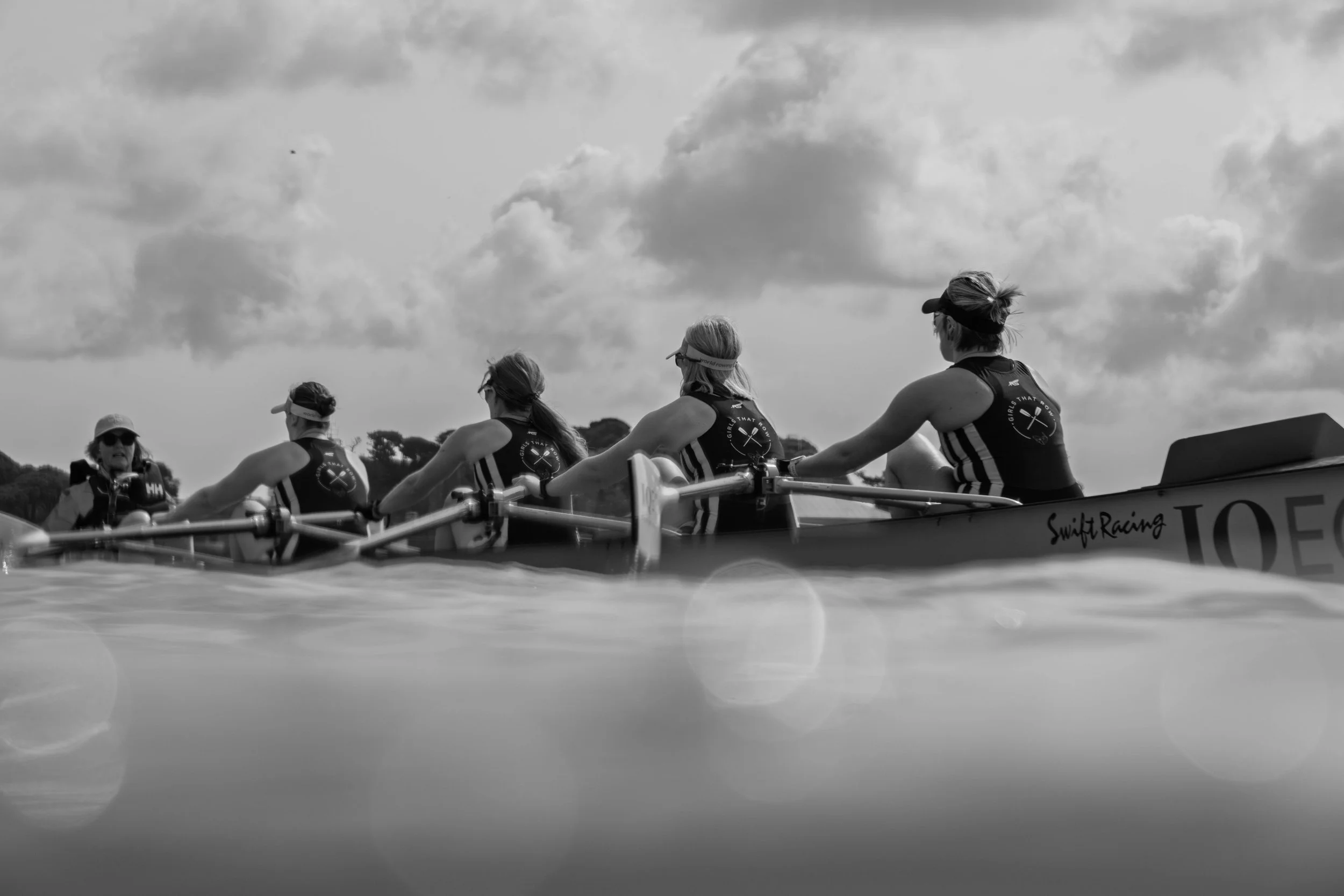 Black and white photo of four women in athletic gear rowing together in a boat under cloudy sky.