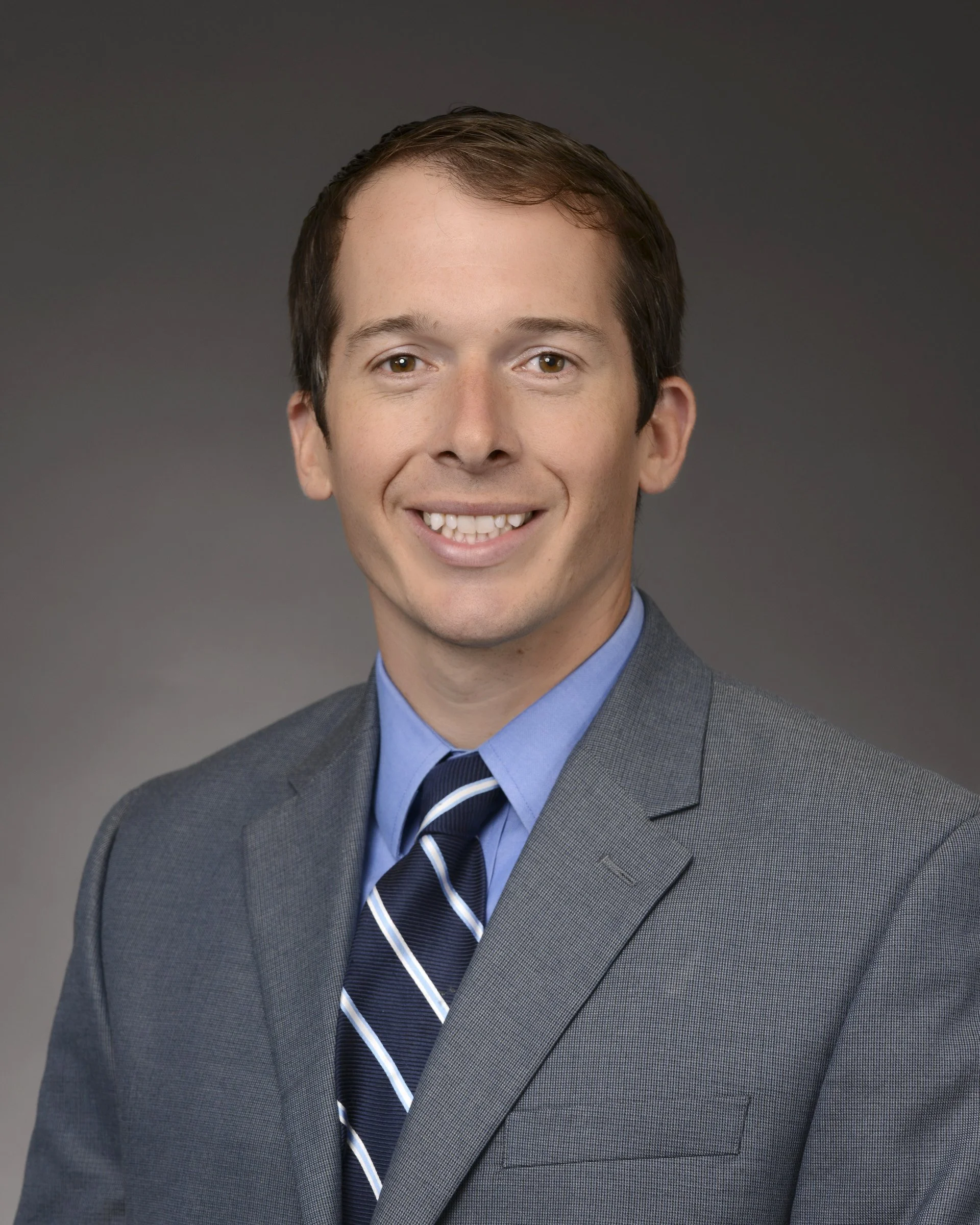 Man wearing a gray suit, blue shirt, and striped tie, smiling against a dark background.