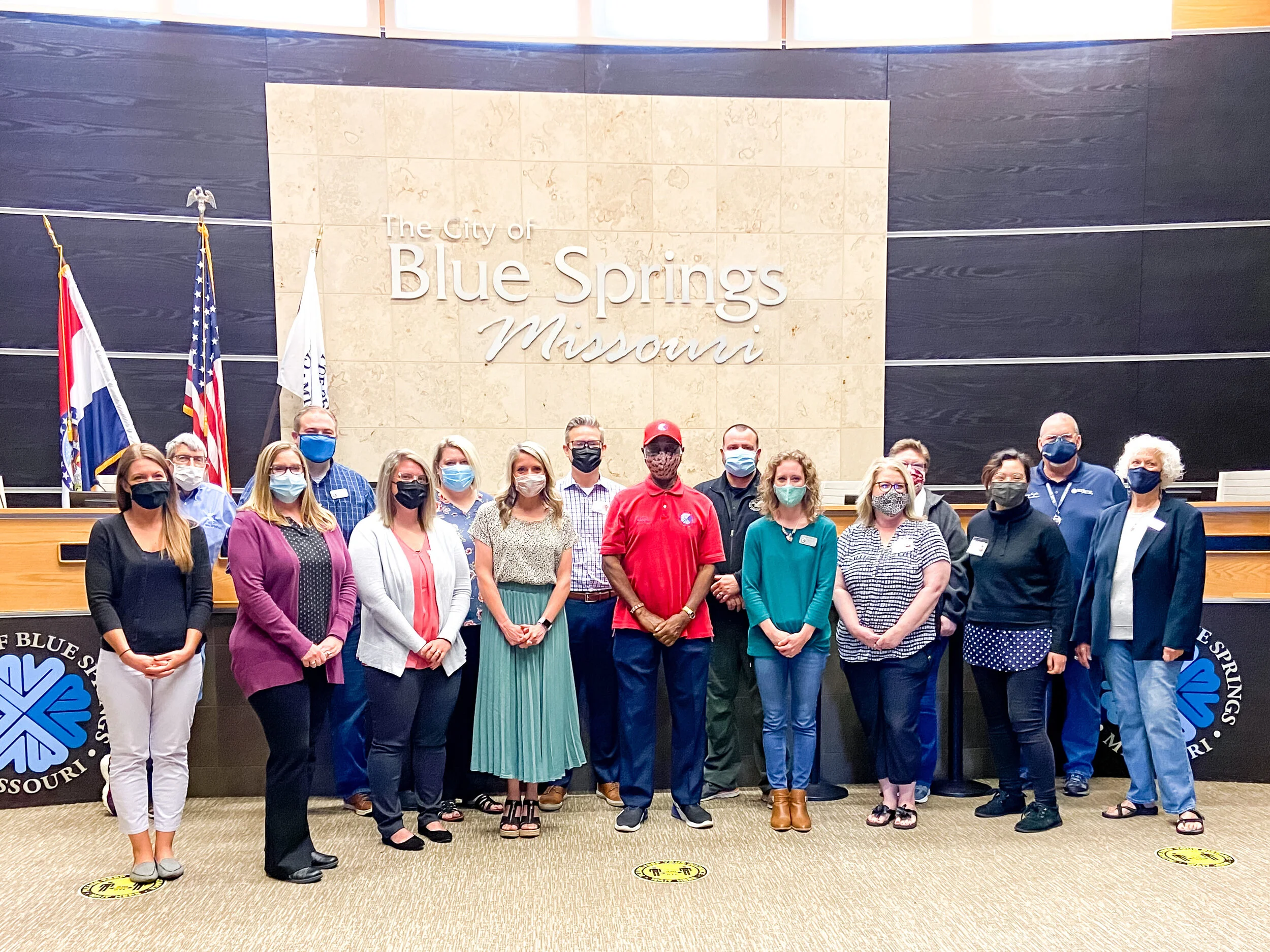 Group of people wearing face masks standing in the City Council chamber of Blue Springs, Missouri. They are in front of a wall with the city's name and state inscribed, along with flags displayed to the side. The group consists of individuals of diverse genders and attire, posing for a formal photograph.