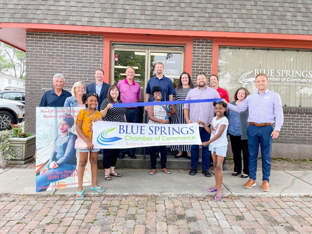 Group of people participating in a ribbon-cutting event for Blue Springs Chamber of Commerce outside a brick building, with a banner and poster displayed.