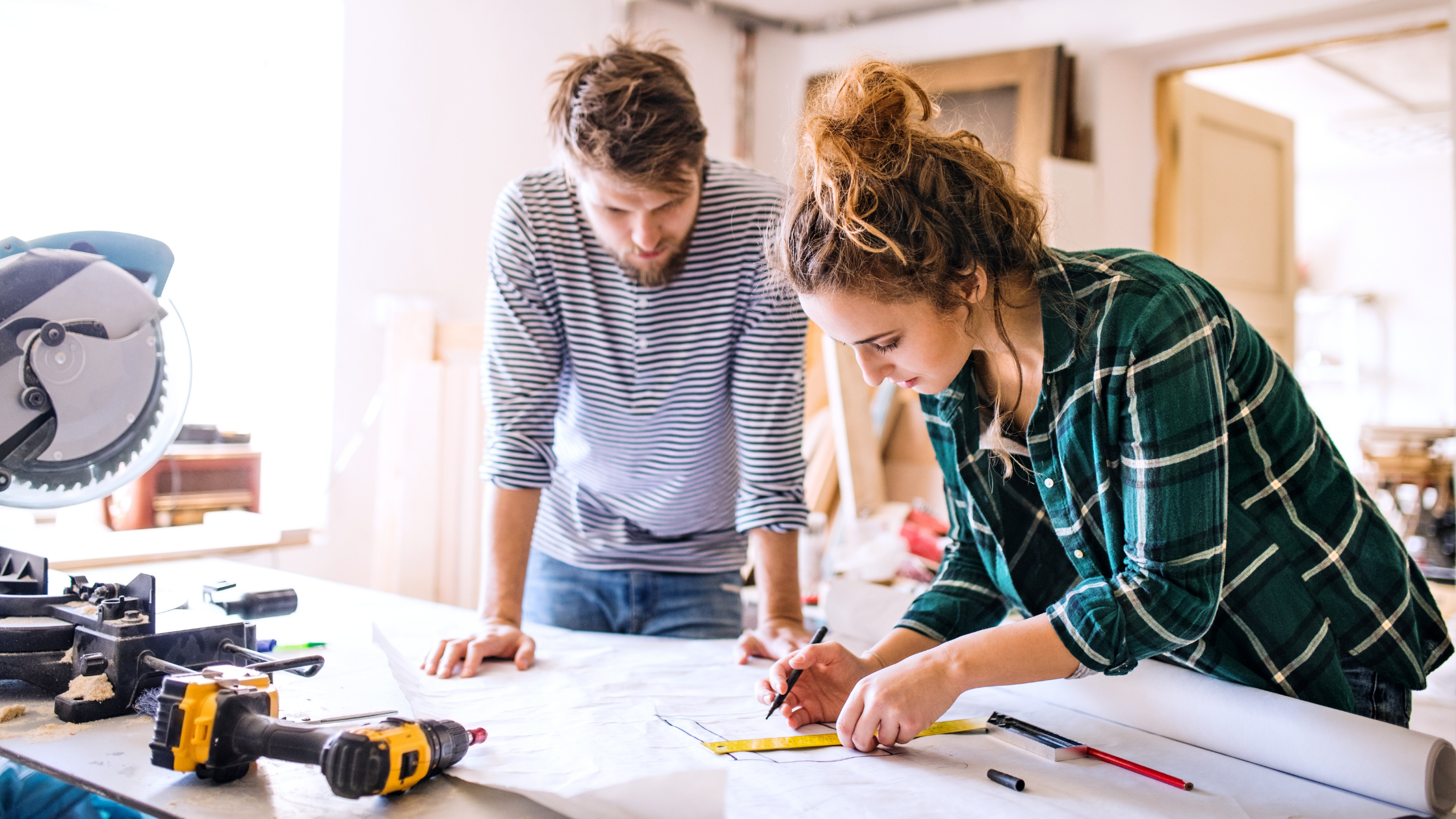 Two people working on a woodwork project in a workshop, using a measuring tape on a blueprint, with tools and a saw on the table.