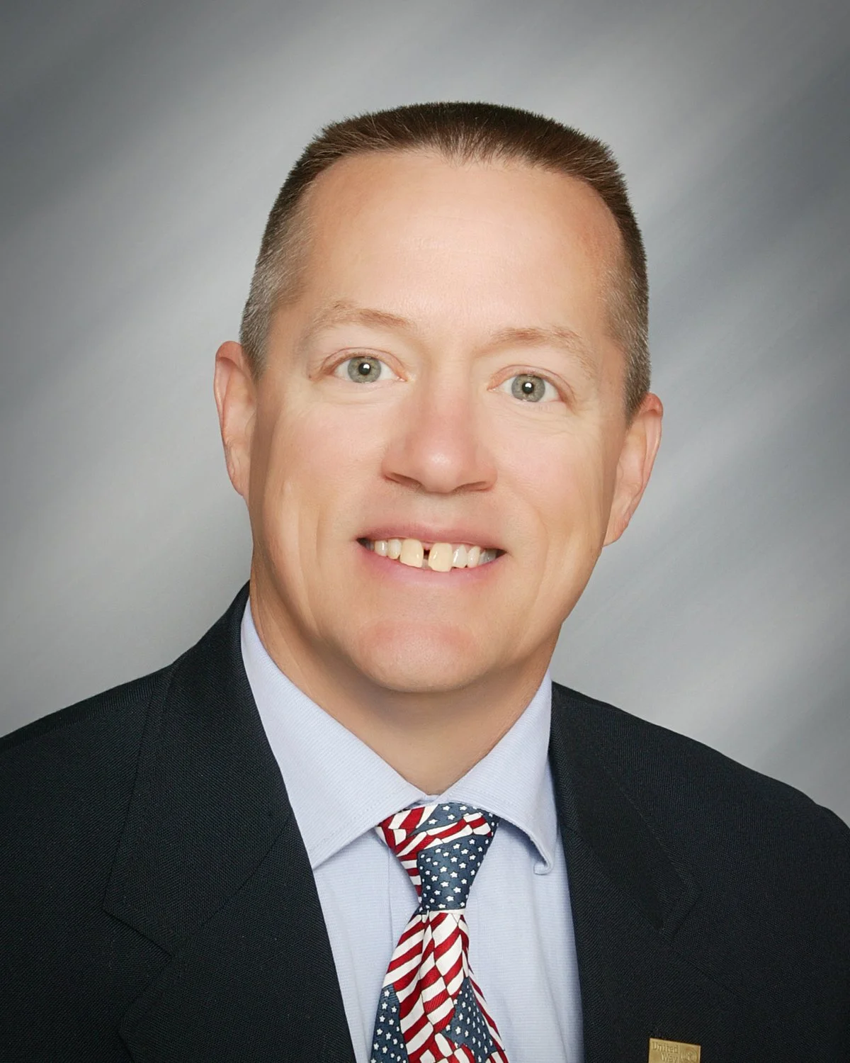 Man in a suit with American flag tie and pin, smiling in portrait.
