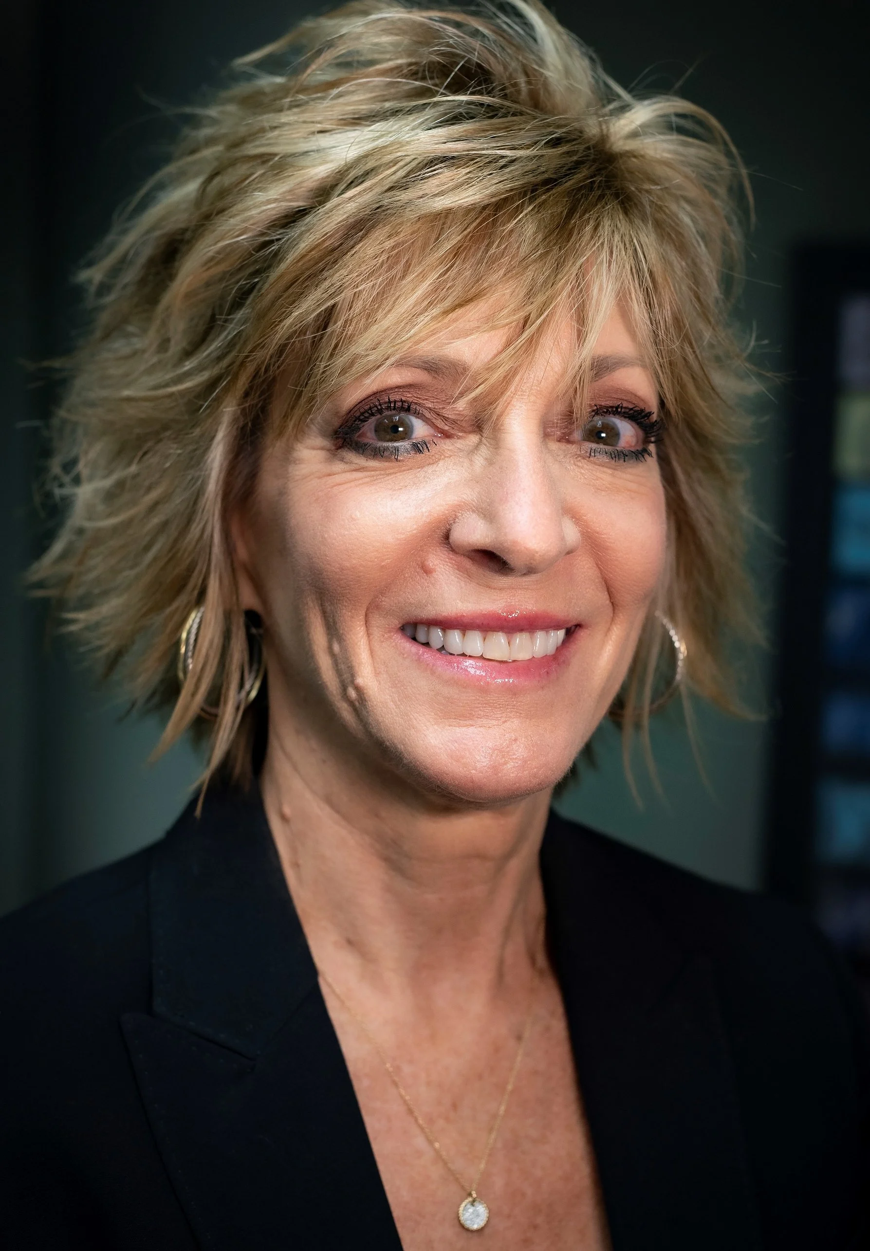 Smiling person against a gray background wearing a purple top and multi-colored striped jacket with short hair.