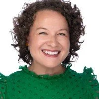 Person with curly hair wearing glasses, smiling, standing in front of a brick wall, dressed in a leopard print top.