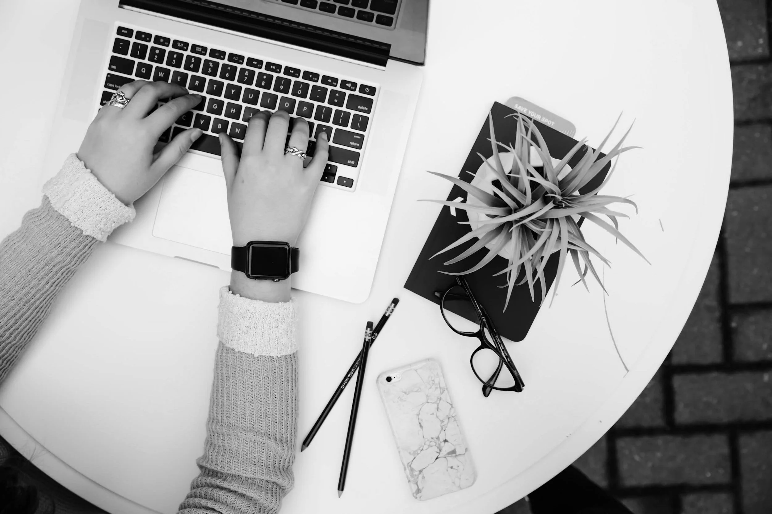 A person using a laptop at a white round table, with a watch on their left wrist and rings on their fingers. The table also has a phone with a marble case, two pencils, a pair of glasses, a closed book, and a potted plant.