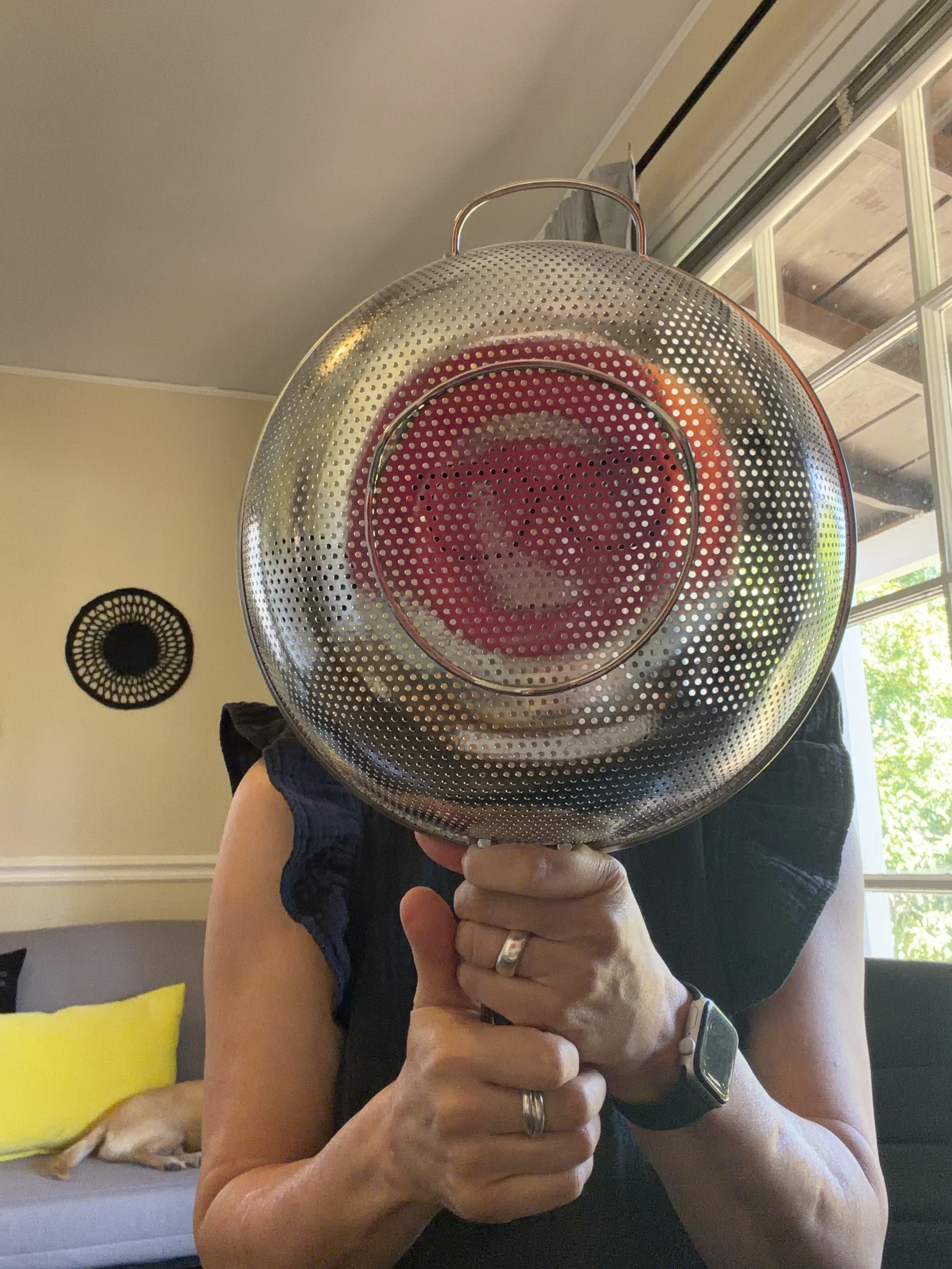 Person taking a selfie using a stainless steel pot as a mirror, holding it in front of their face while wearing a ring, a watch, and a dark sleeveless top, with a window and wall decor visible in the background.