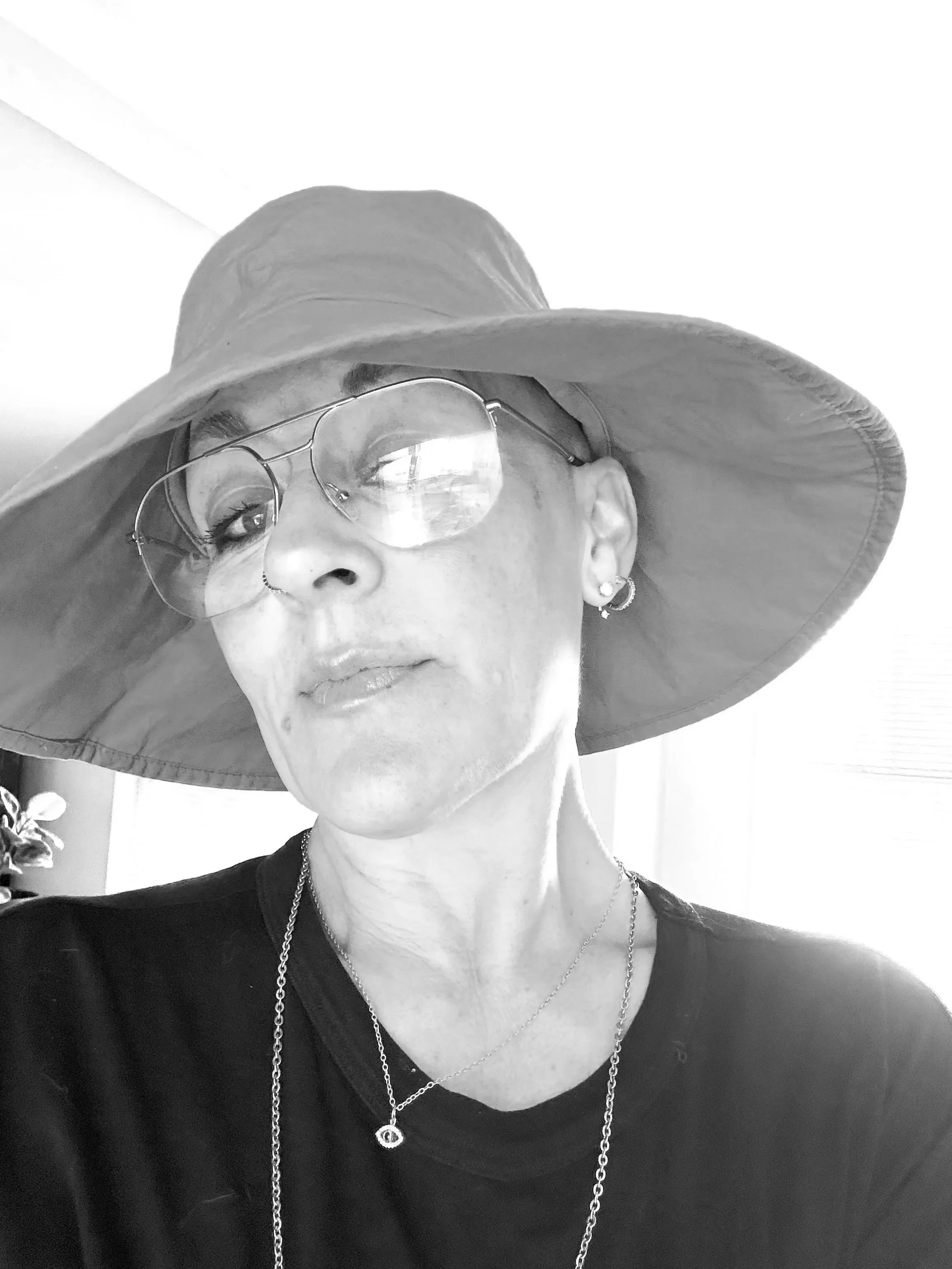 A woman wearing a large brimmed hat, glasses, earrings, a necklace, and a dark top, looking slightly to the side.