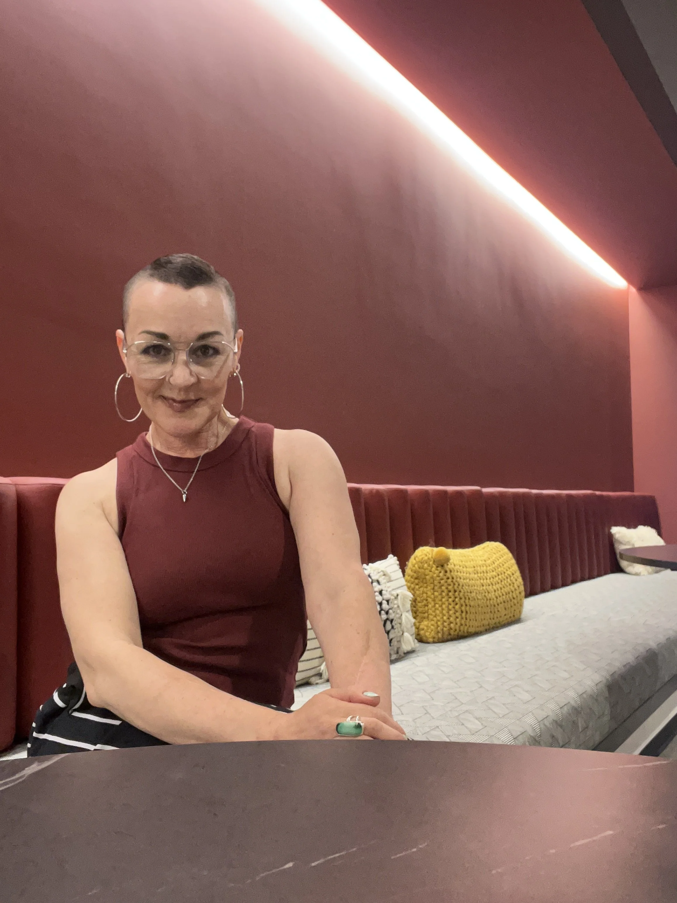 A woman with short hair, glasses, and hoop earrings sitting at a table in a room with maroon walls and modern lighting, wearing a sleeveless maroon top and a necklace, with a yellow knitted purse and decorative pillows on a bed behind her.